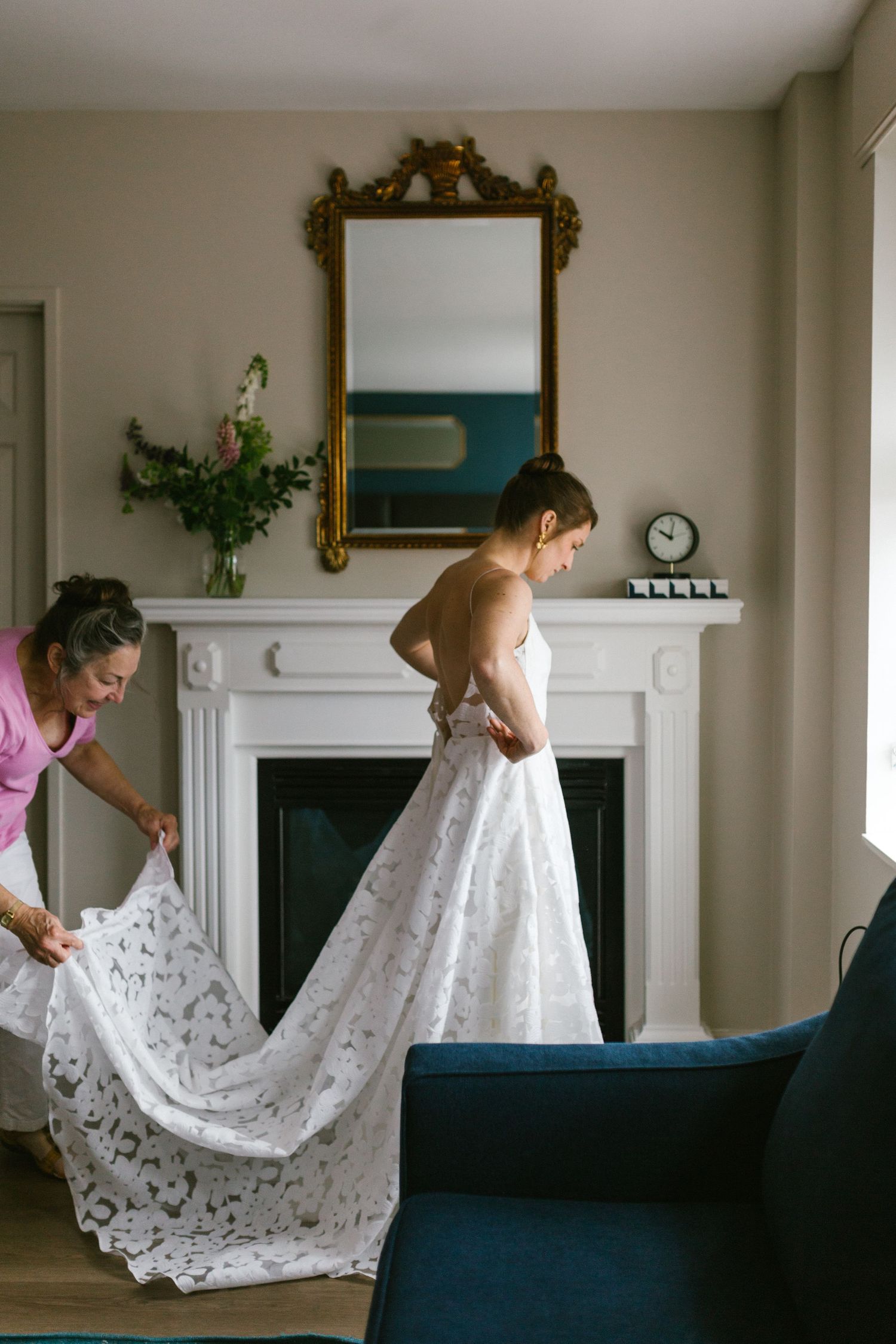 A delicate lace wedding gown being fitted in an elegant room with a white fireplace and ornate gold-framed mirror.