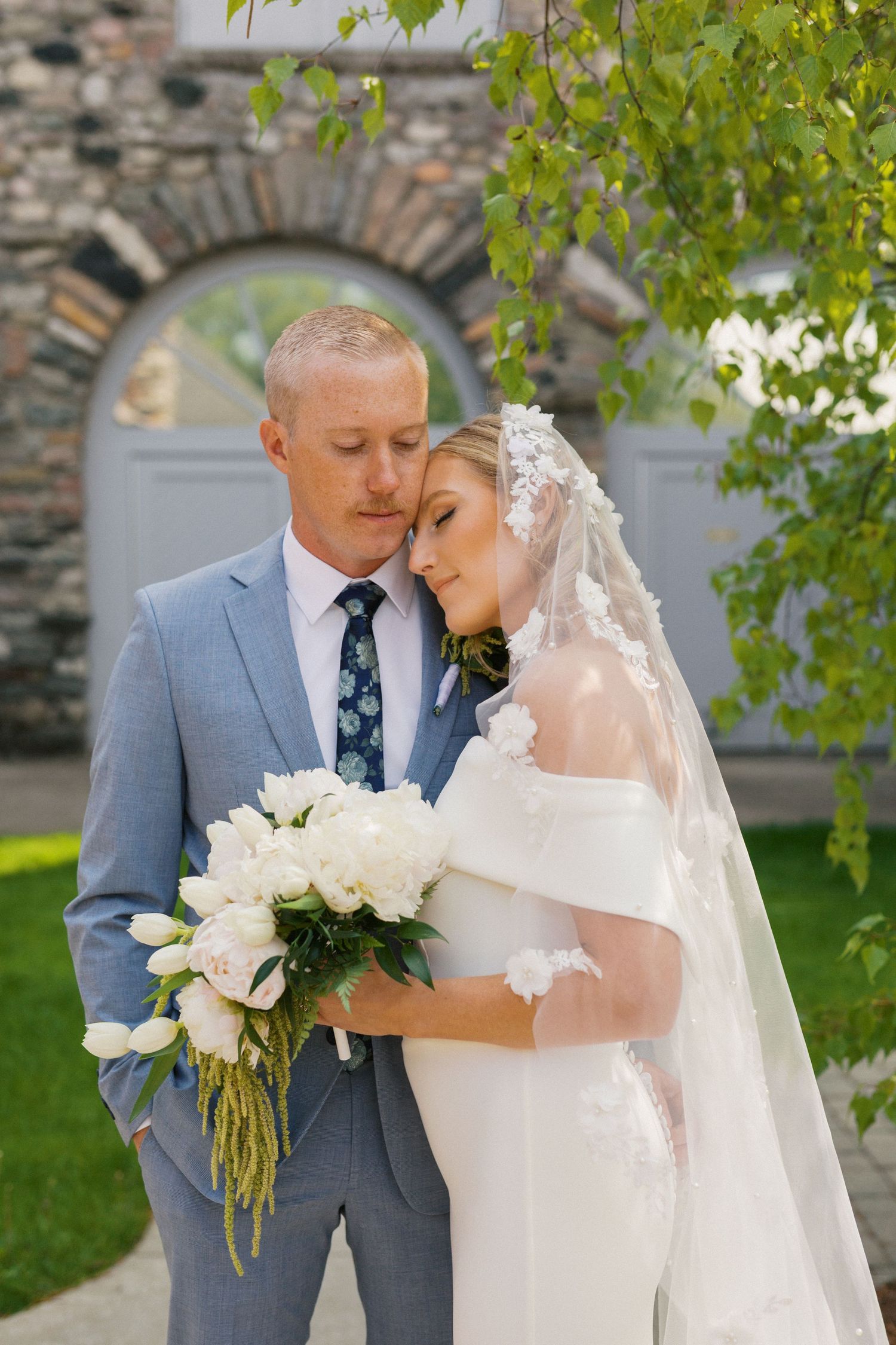 Romantic wedding couple shares intimate moments against stone wall backdrop while holding white floral bouquet.
