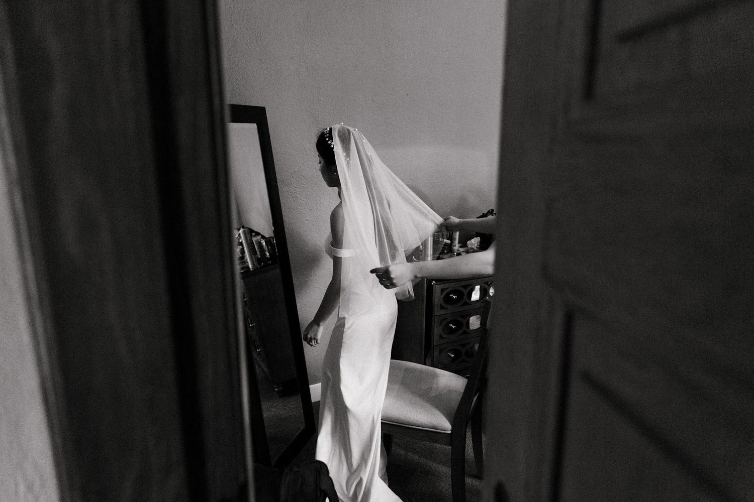 Black and white photo of a bride holding her wedding veil in a sunlit room with a mirror and chair.