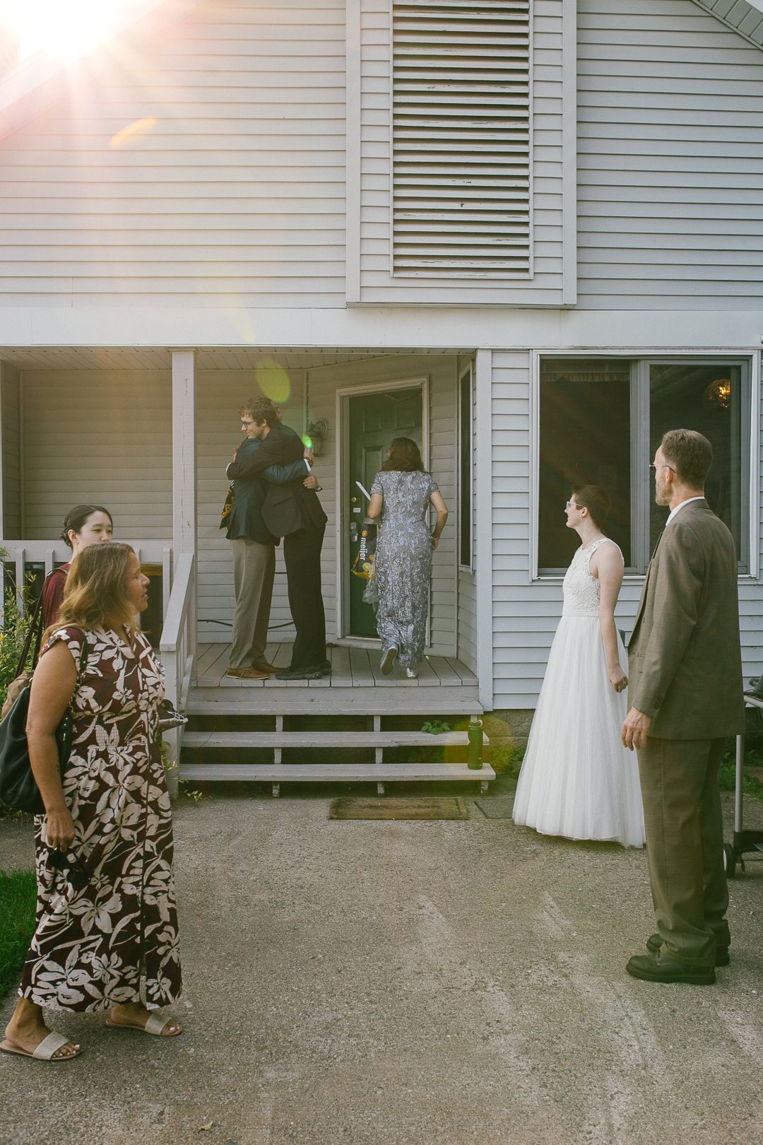 Wedding guests gather outside a white clapboard house with formal attire and a wedding dress visible.