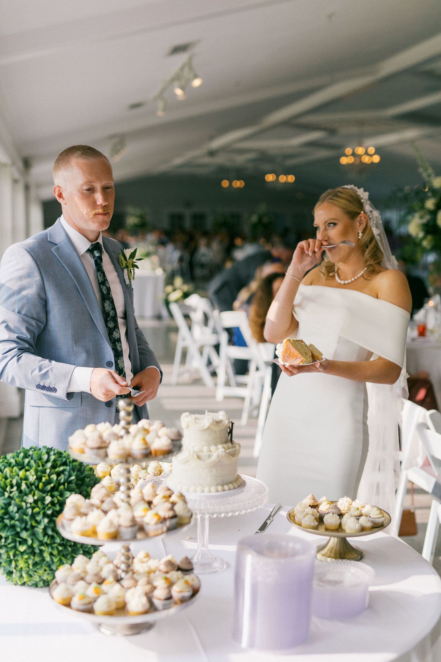 Wedding dessert table with elegant white cake and cupcakes displayed under crystal chandeliers at elegant reception.