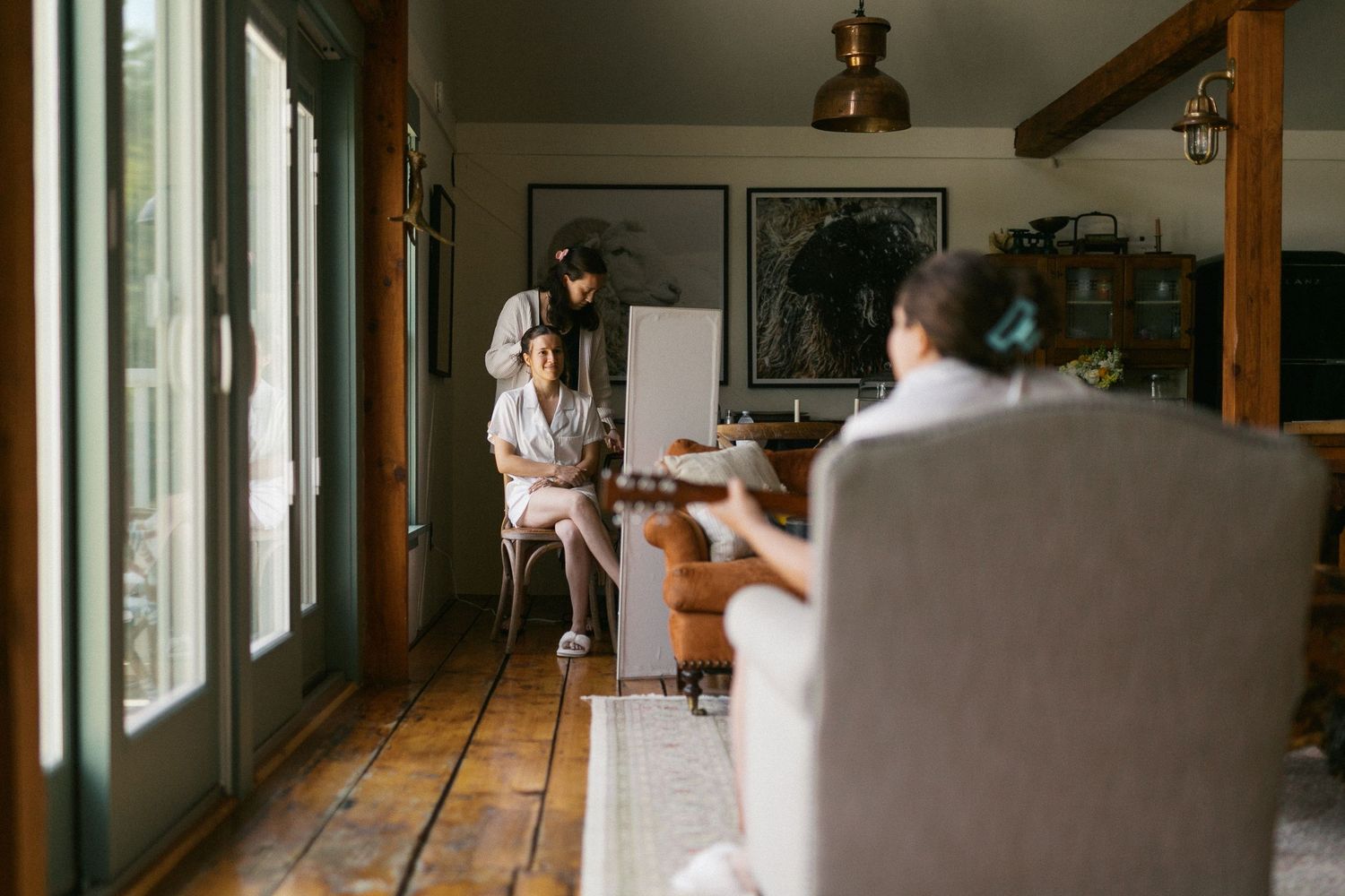 Three people relaxing in a cozy rustic living room with hardwood floors, large windows, and framed artwork on the walls.