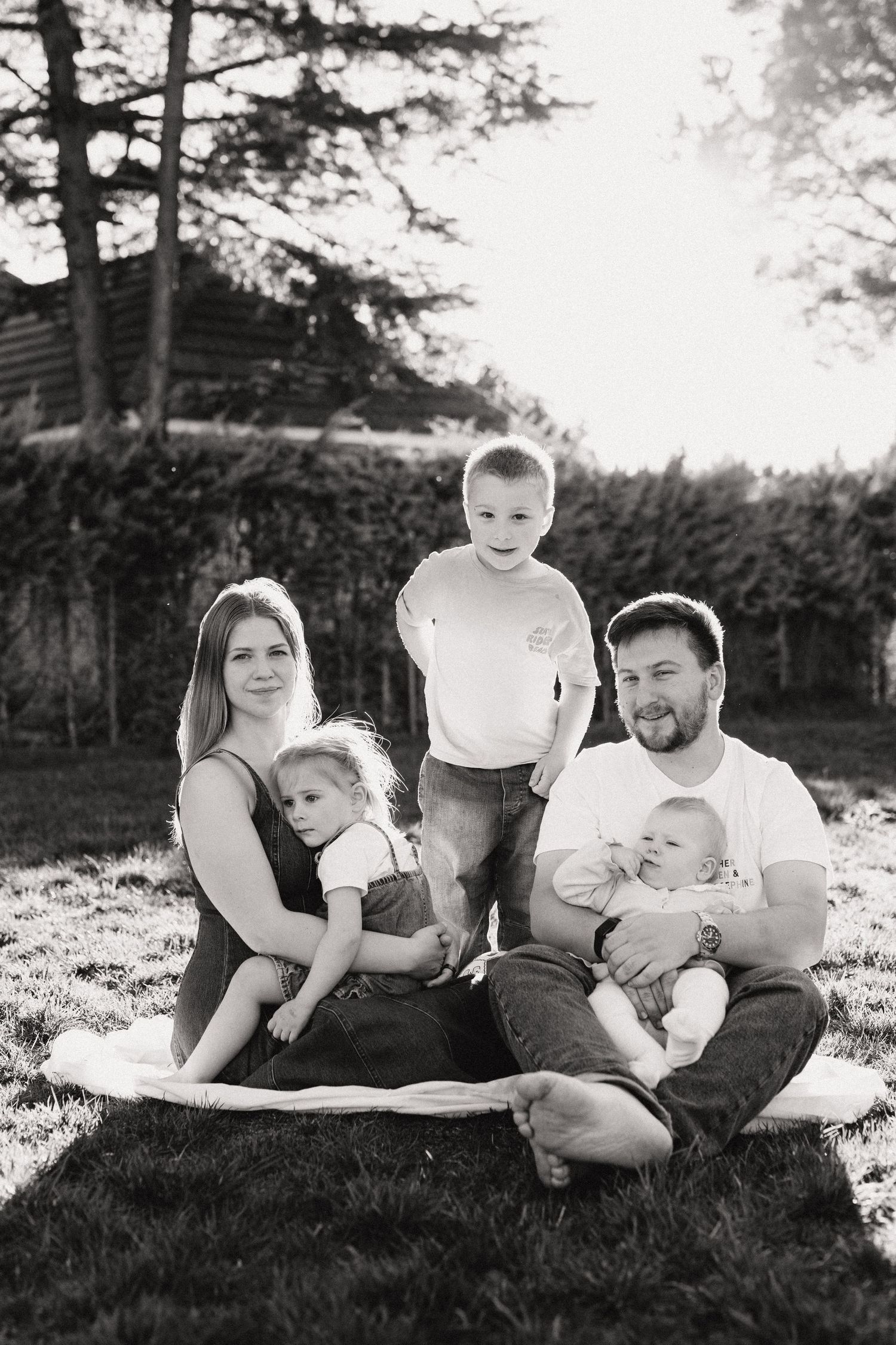 Black and white family portrait of parents with three young children sitting on grass in a sunny backyard.