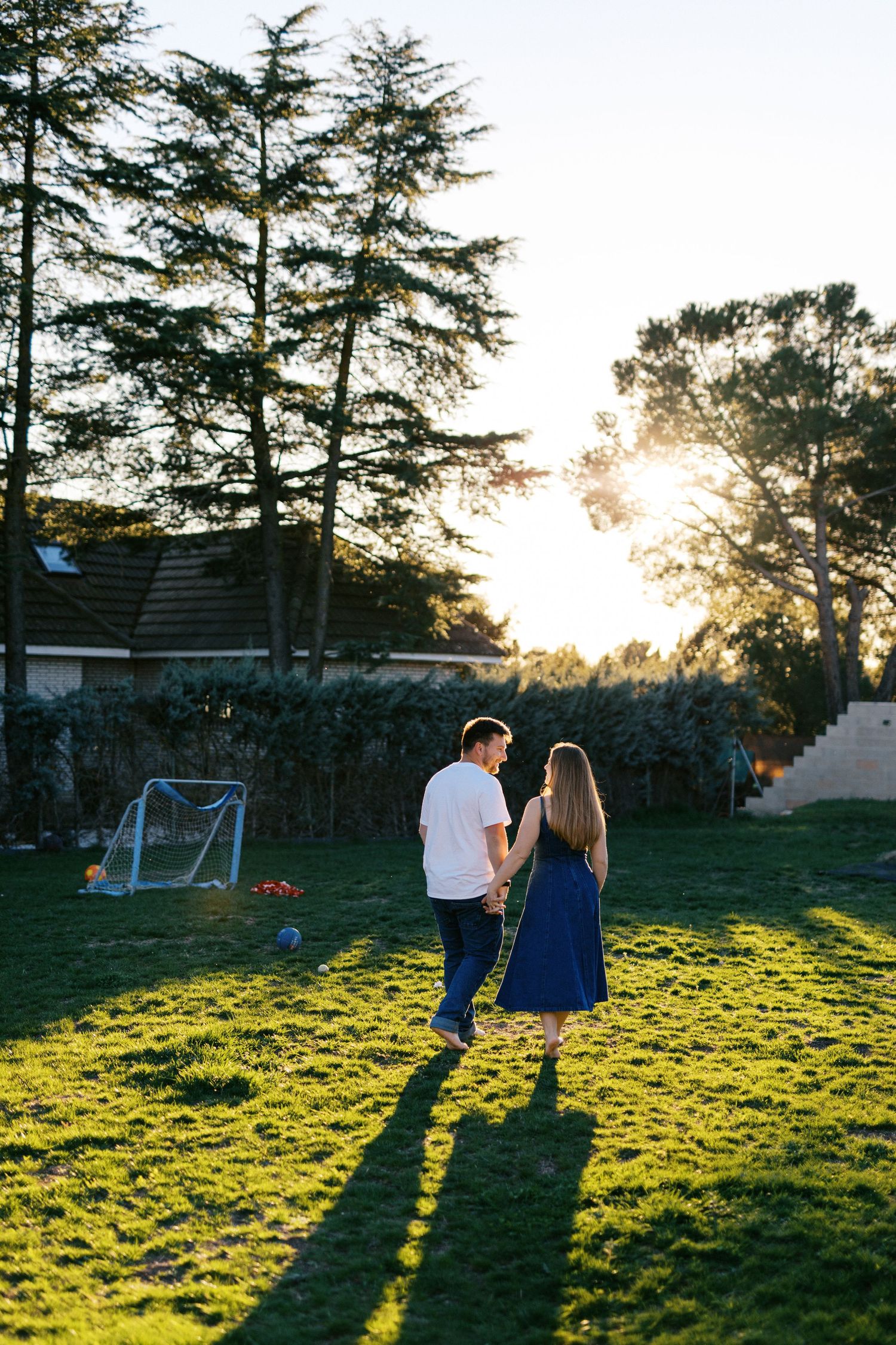 Couple holding hands walking barefoot on green grass in backyard at golden sunset.