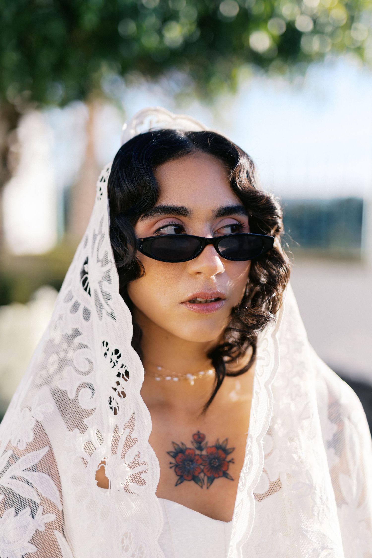 Edgy bride with black sunglasses, lace mantilla veil, and floral chest tattoo poses outdoors in sunlight.