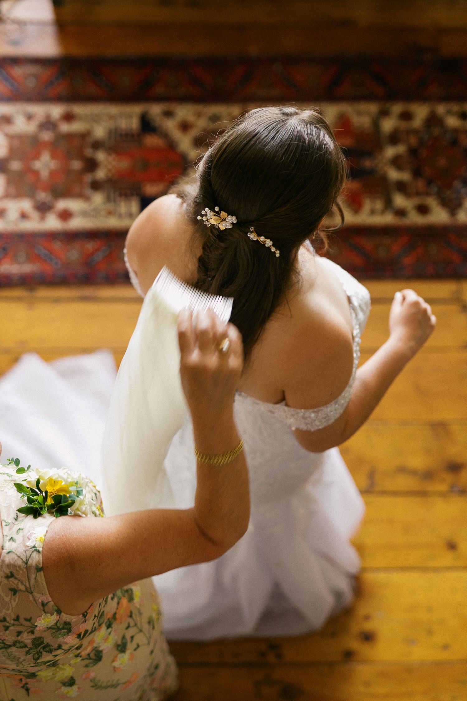 Bride in white lace off-shoulder gown gets veil adjusted, hair adorned with gold pins, on wooden floor.