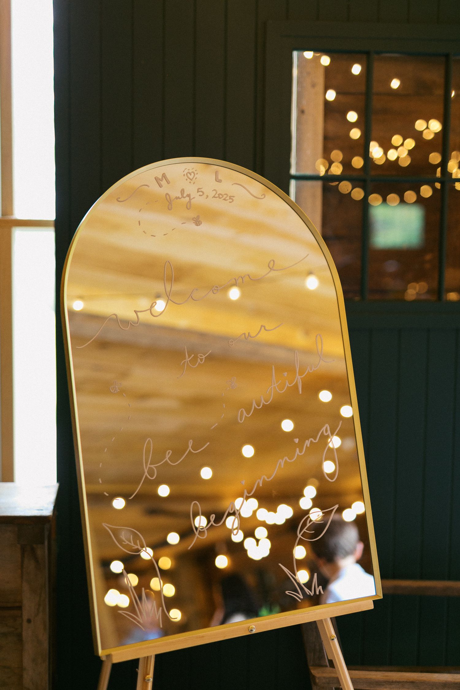 Gold arched mirror on easel with elegant calligraphy script, reflecting warm bokeh string lights at a wedding venue.