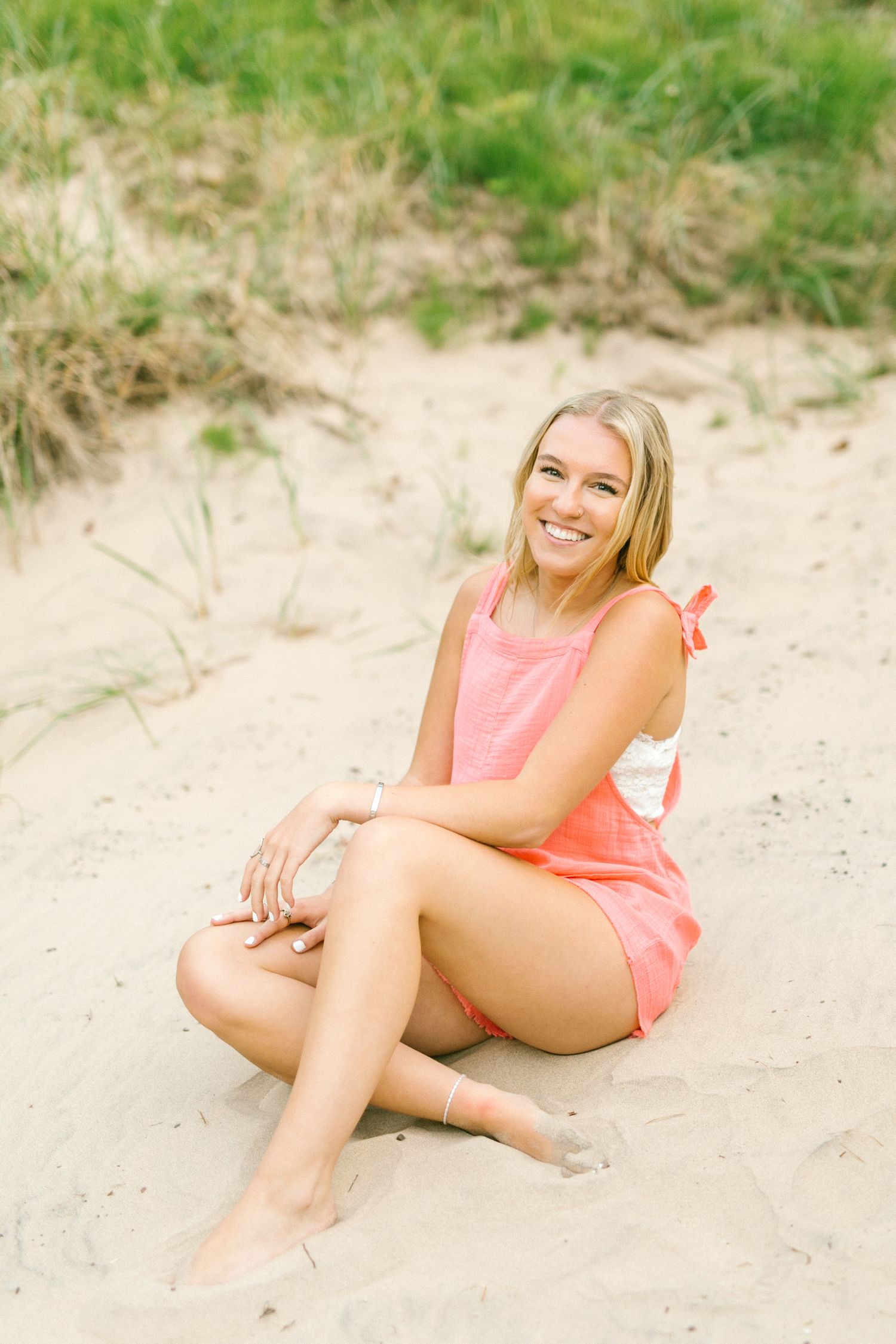 Smiling blonde woman in pink romper sitting on sandy beach dunes.