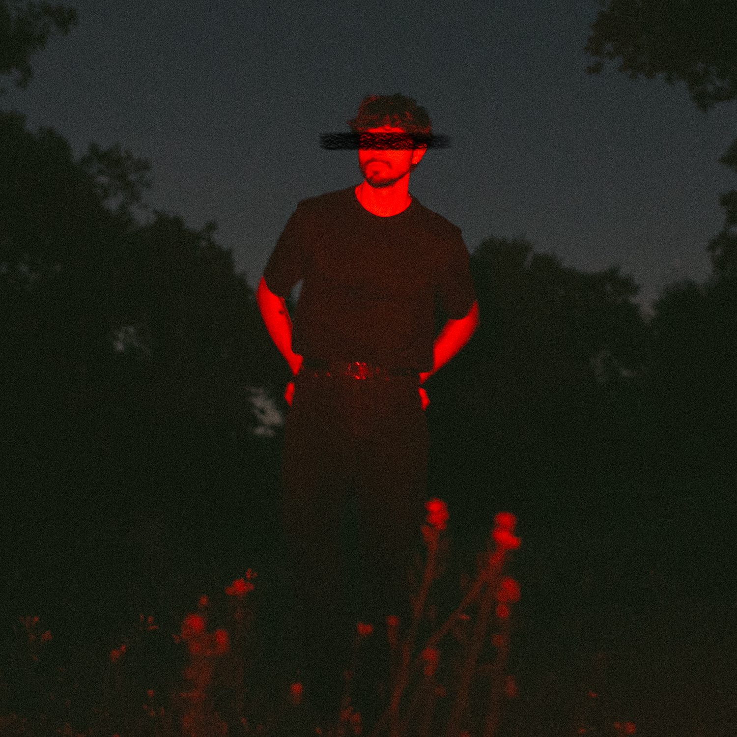 A mysterious figure stands in darkness illuminated by dramatic red light wearing a wide-brim hat outdoors at night.