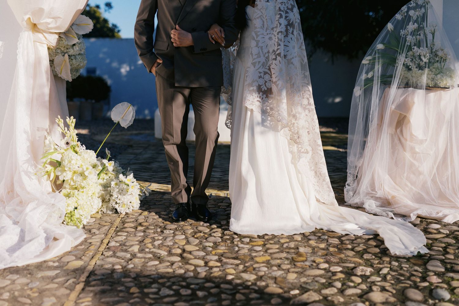 Close-up of bride and groom walking on cobblestones surrounded by white floral draping and arrangements.