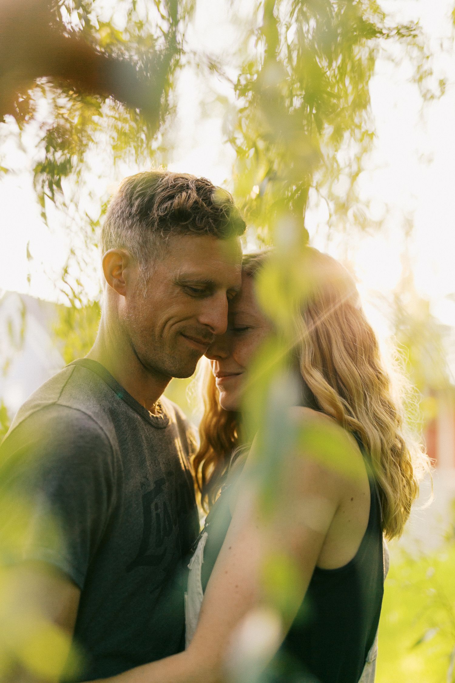 Couple sharing an intimate forehead touch framed by sun-drenched green leaves in warm golden light.