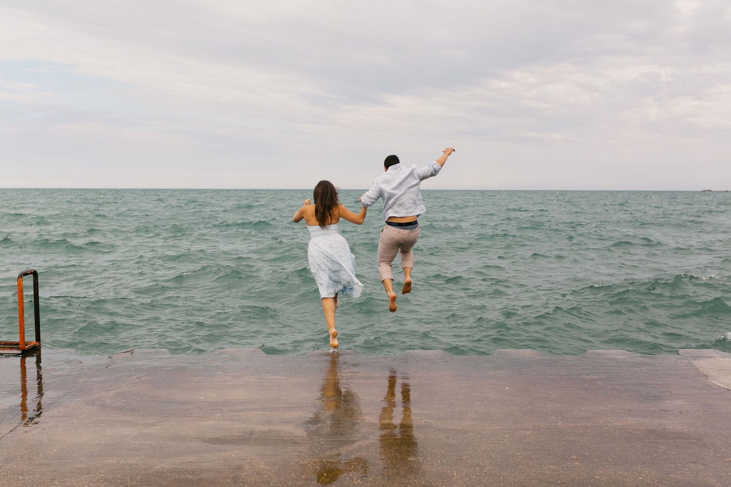 Couple joyfully jumping into Lake Michigan from a pier on an overcast day.