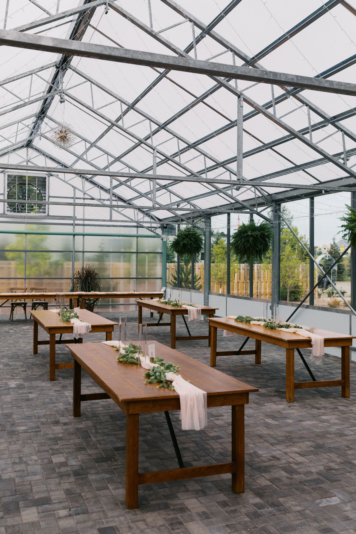 Elegant greenhouse venue with wooden farm tables, greenery runners, and hanging plants for a rustic wedding reception.