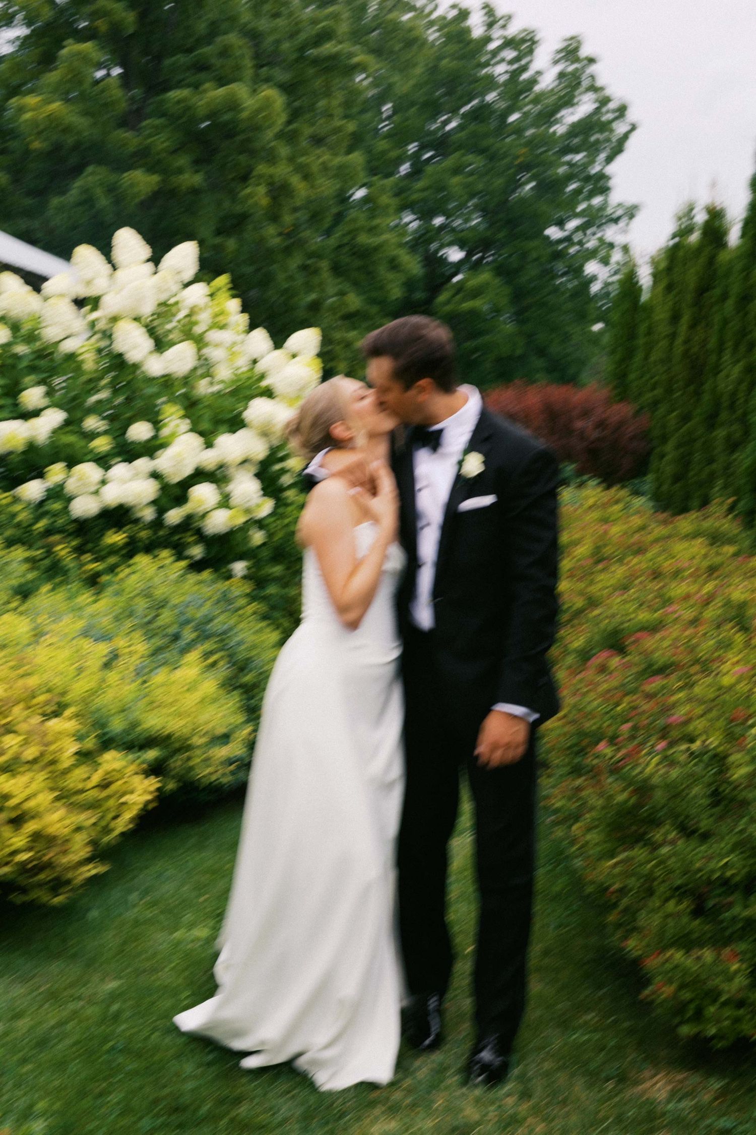 A romantic wedding couple shares a tender moment in a lush garden setting with white hydrangea bushes in bloom.