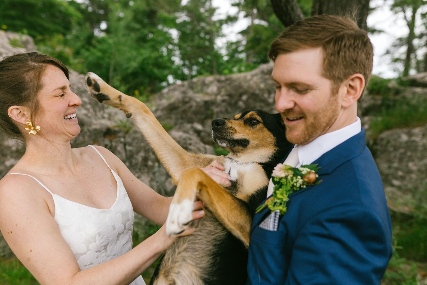 A joyful couple in wedding attire shares a playful moment with their small brown dog against a rocky outdoor backdrop.