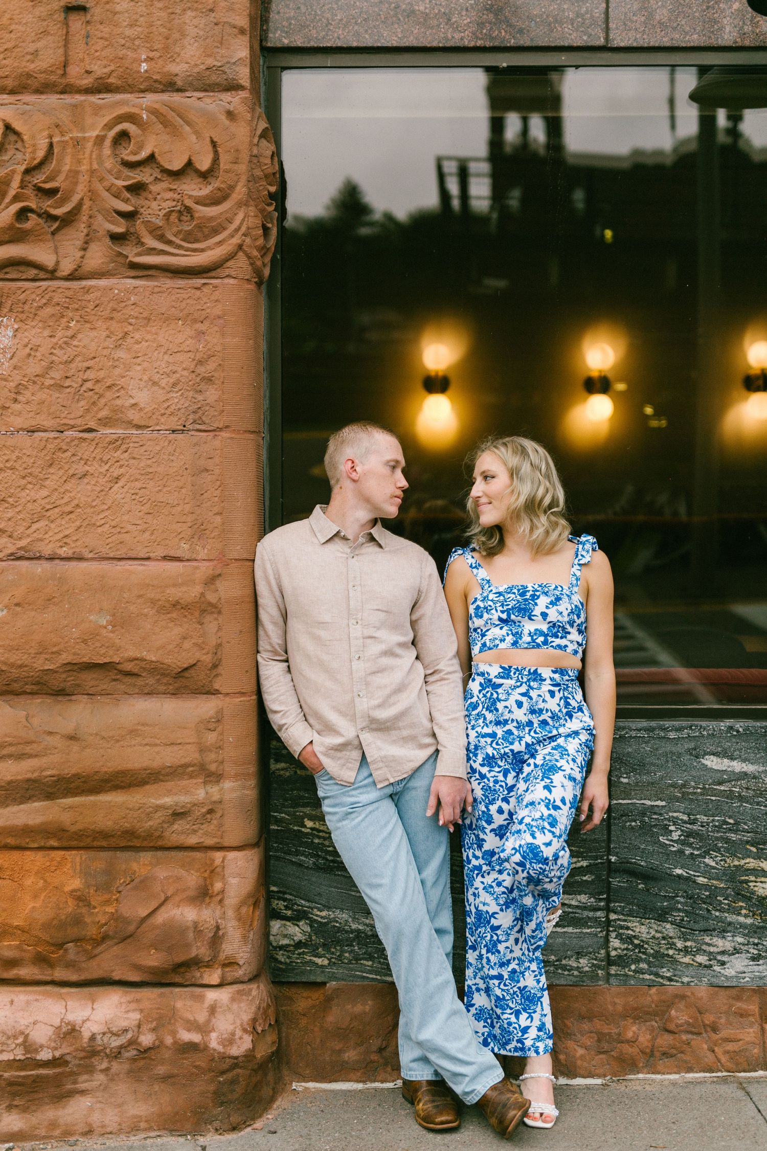A couple leans against a stone wall with warm lighting at dusk, the woman wearing a blue floral outfit and man in beige.