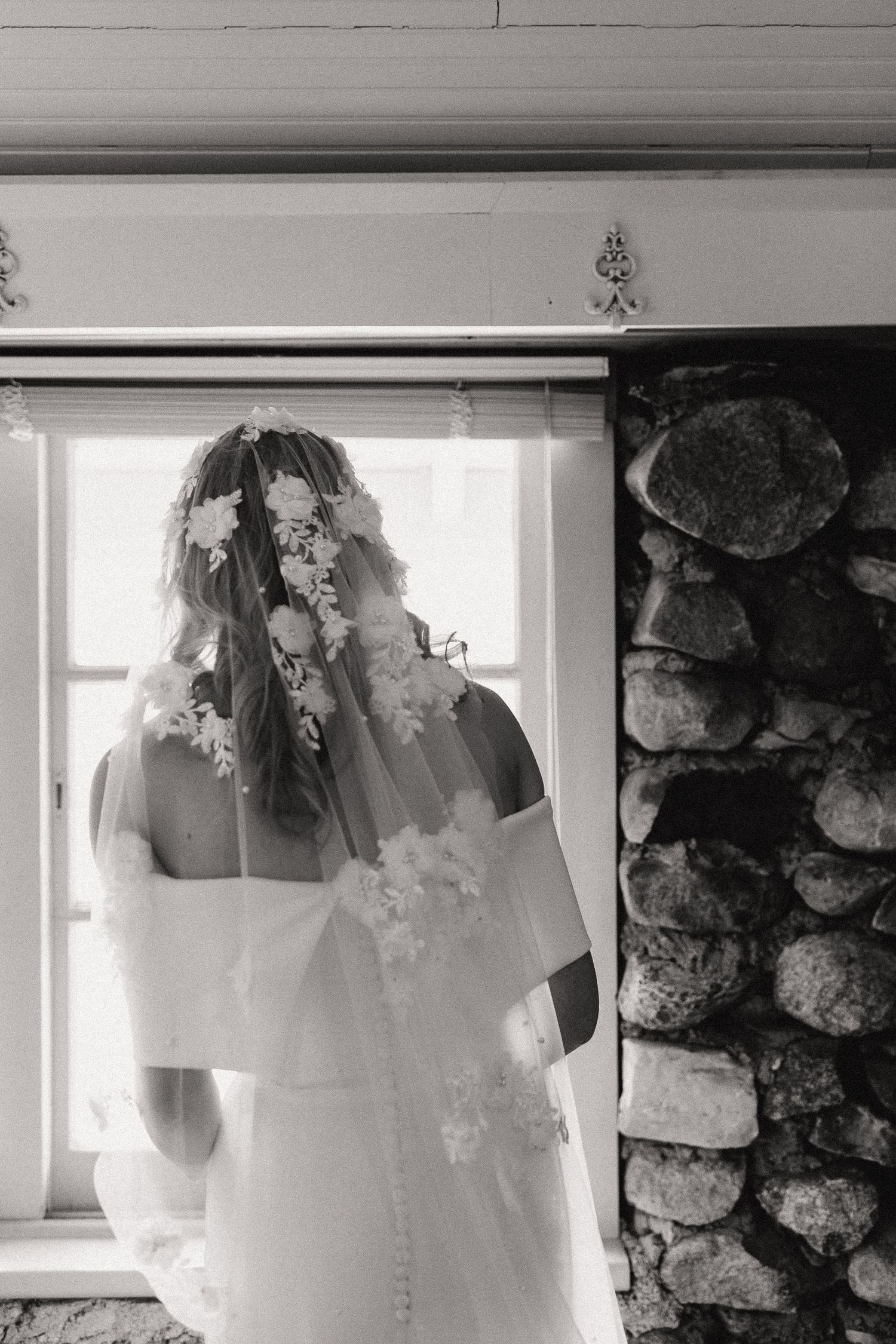 A black and white photo showing a wedding dress and veil near a stone wall and window.