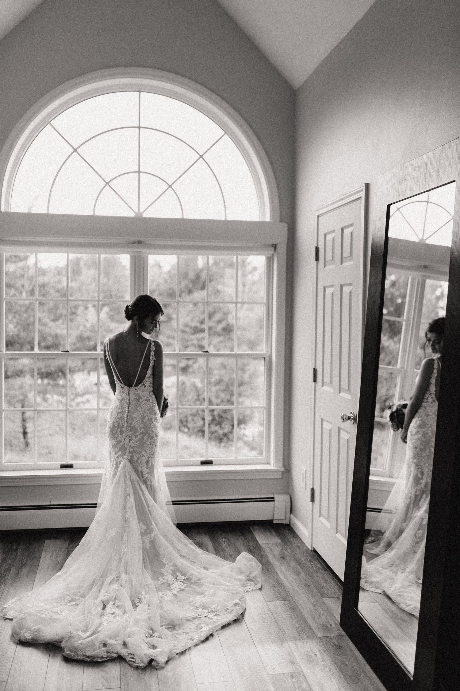 Elegant bride in lace wedding gown stands by arched window with reflection in full-length mirror showing train detail.