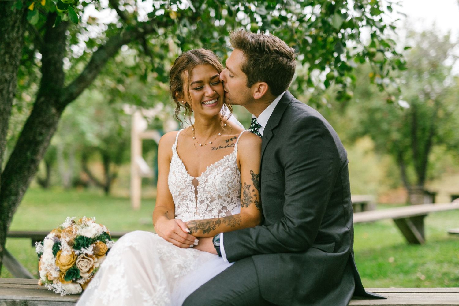 A tender wedding moment captured outdoors with a couple sitting on a bench surrounded by greenery.