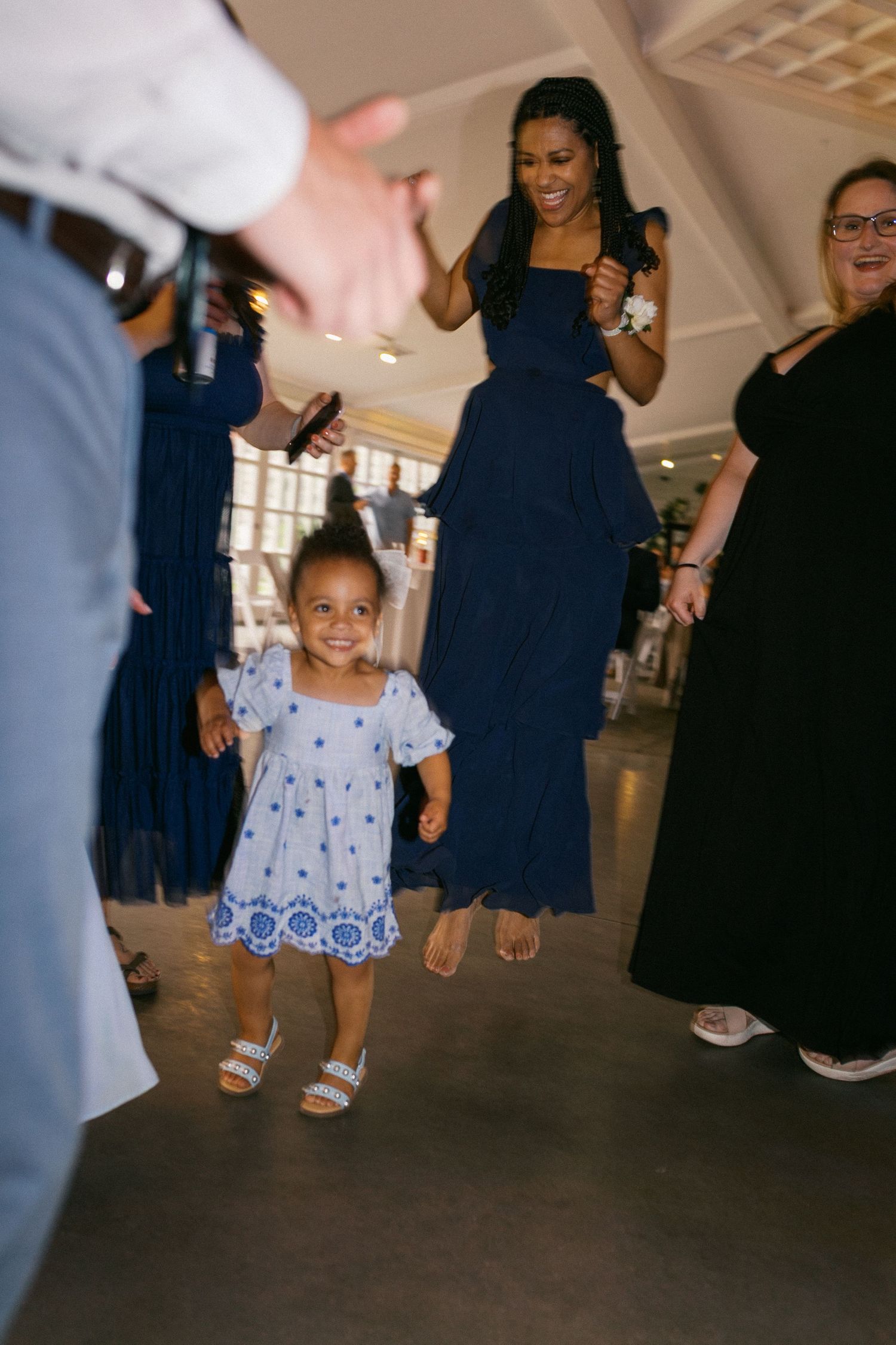 A small child in a blue dress dances at a wedding reception surrounded by guests in navy attire.