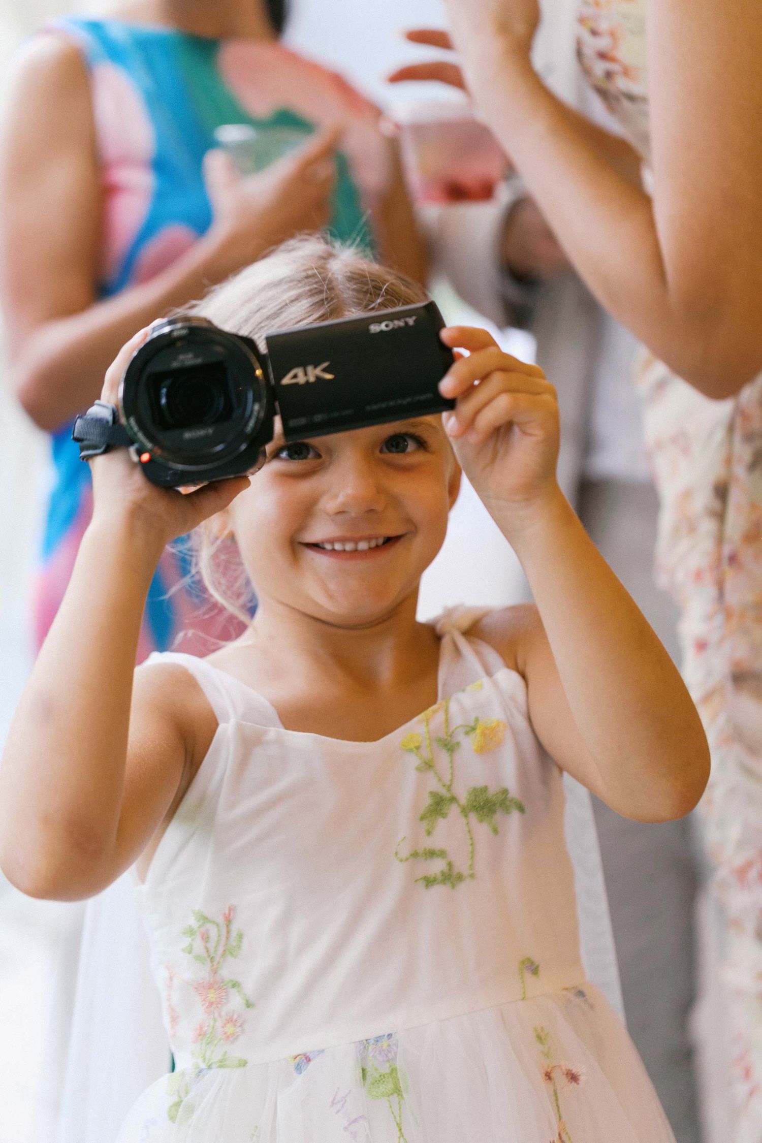A young videographer holds up a Sony 4K camera while wearing a white floral dress.