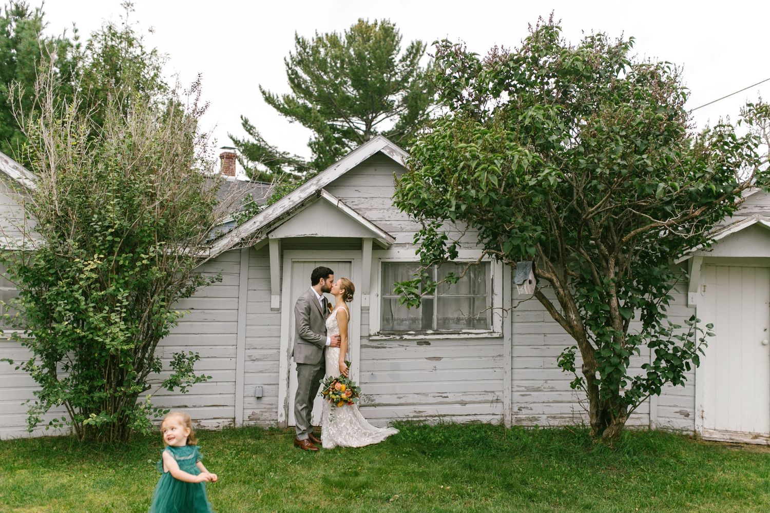 A couple stands in the doorway of a small grey house surrounded by lush green trees on their wedding day.