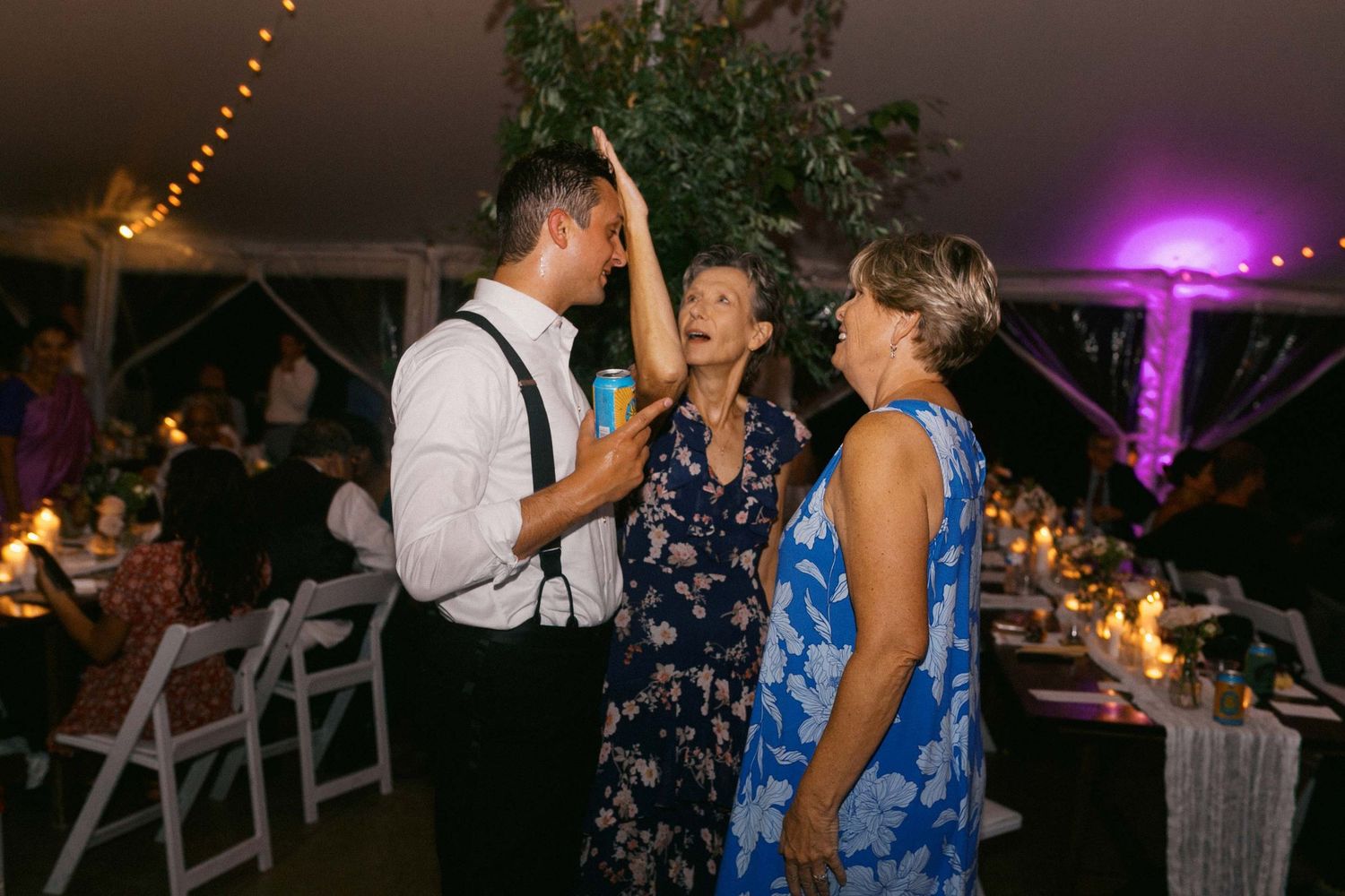Wedding guests dance and celebrate together on an outdoor dance floor illuminated by string lights and purple uplighting.