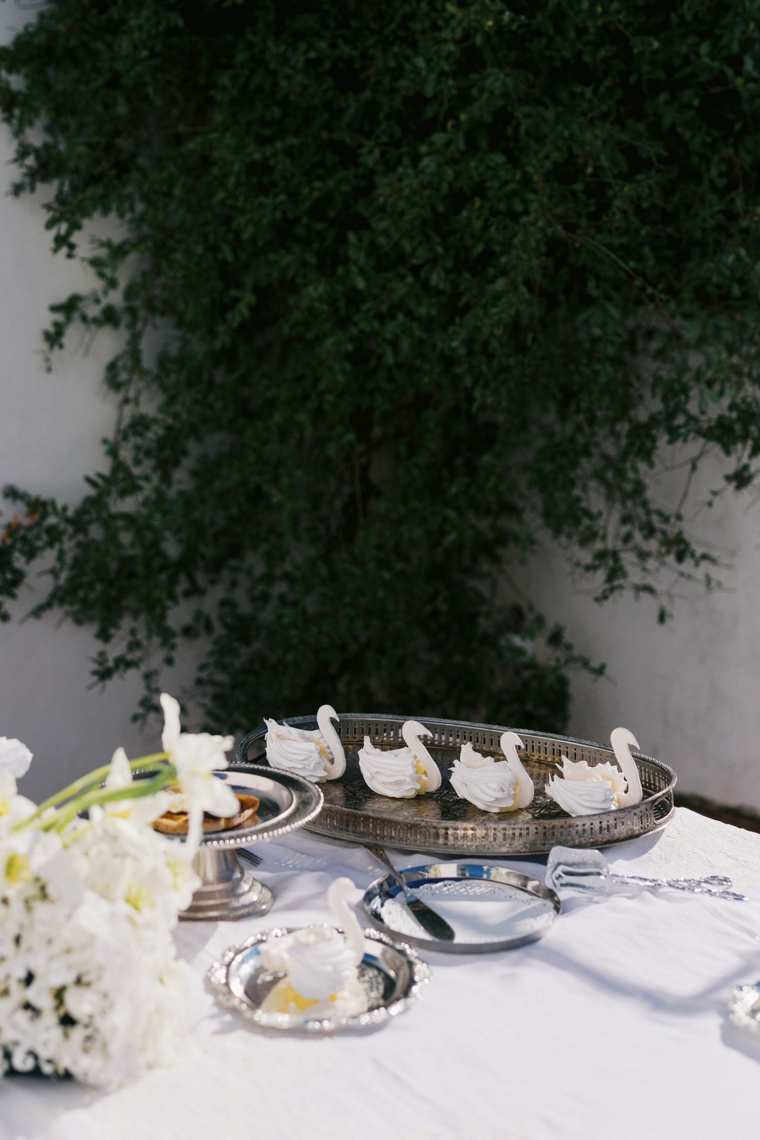 Elegant wedding dessert table with swan-shaped pastries on silver tray, white flowers, and silver accessories outdoors.