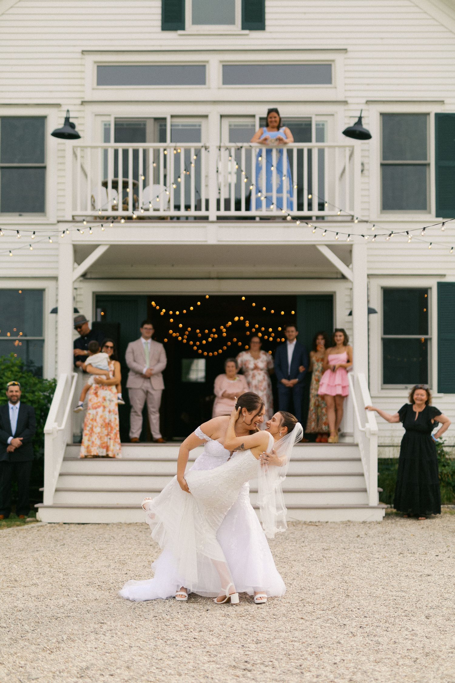 Two brides share a romantic dip kiss during their outdoor wedding reception in front of a white venue.