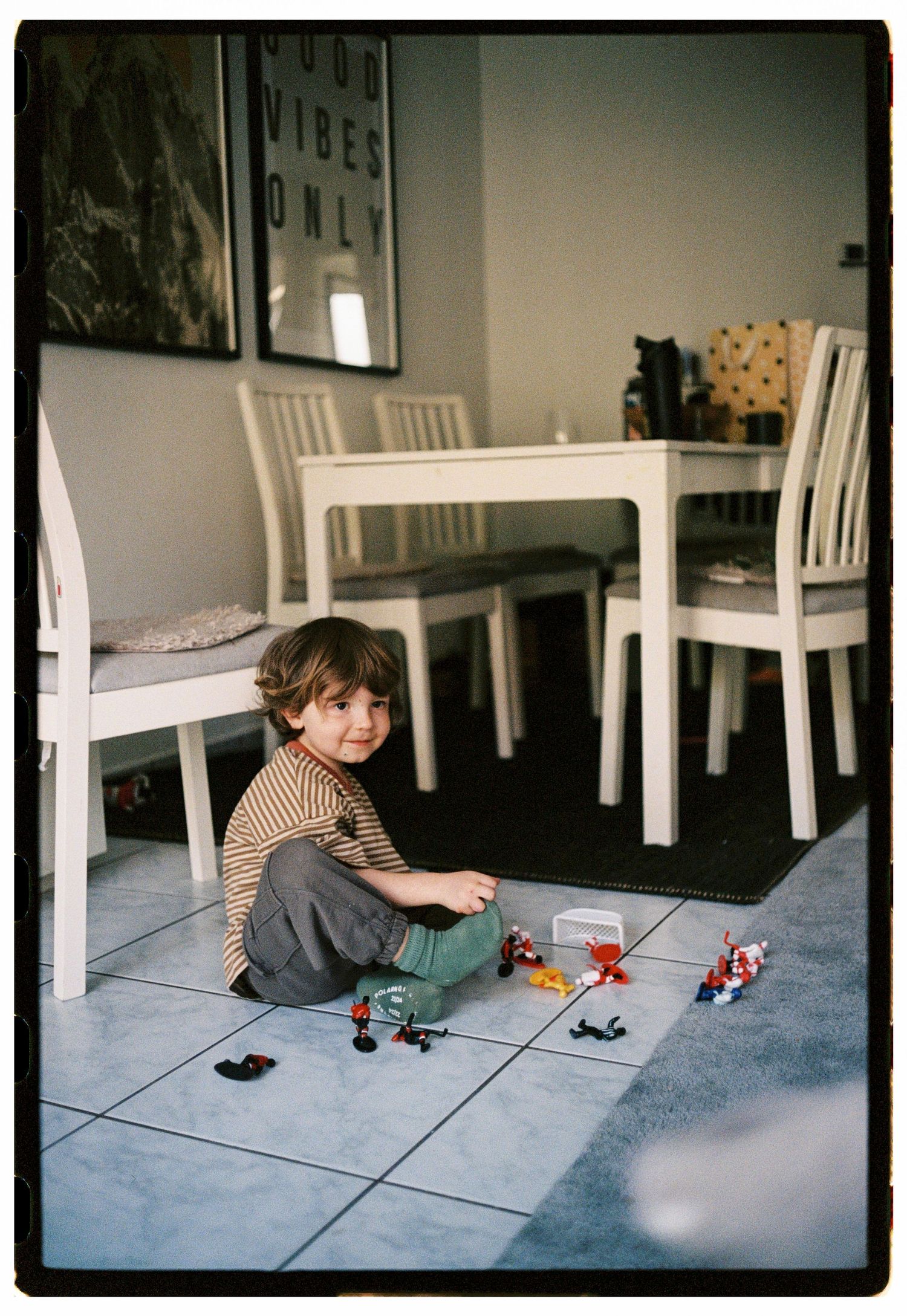 Smiling toddler sitting on tile floor playing with colorful toy figures near white dining chairs.