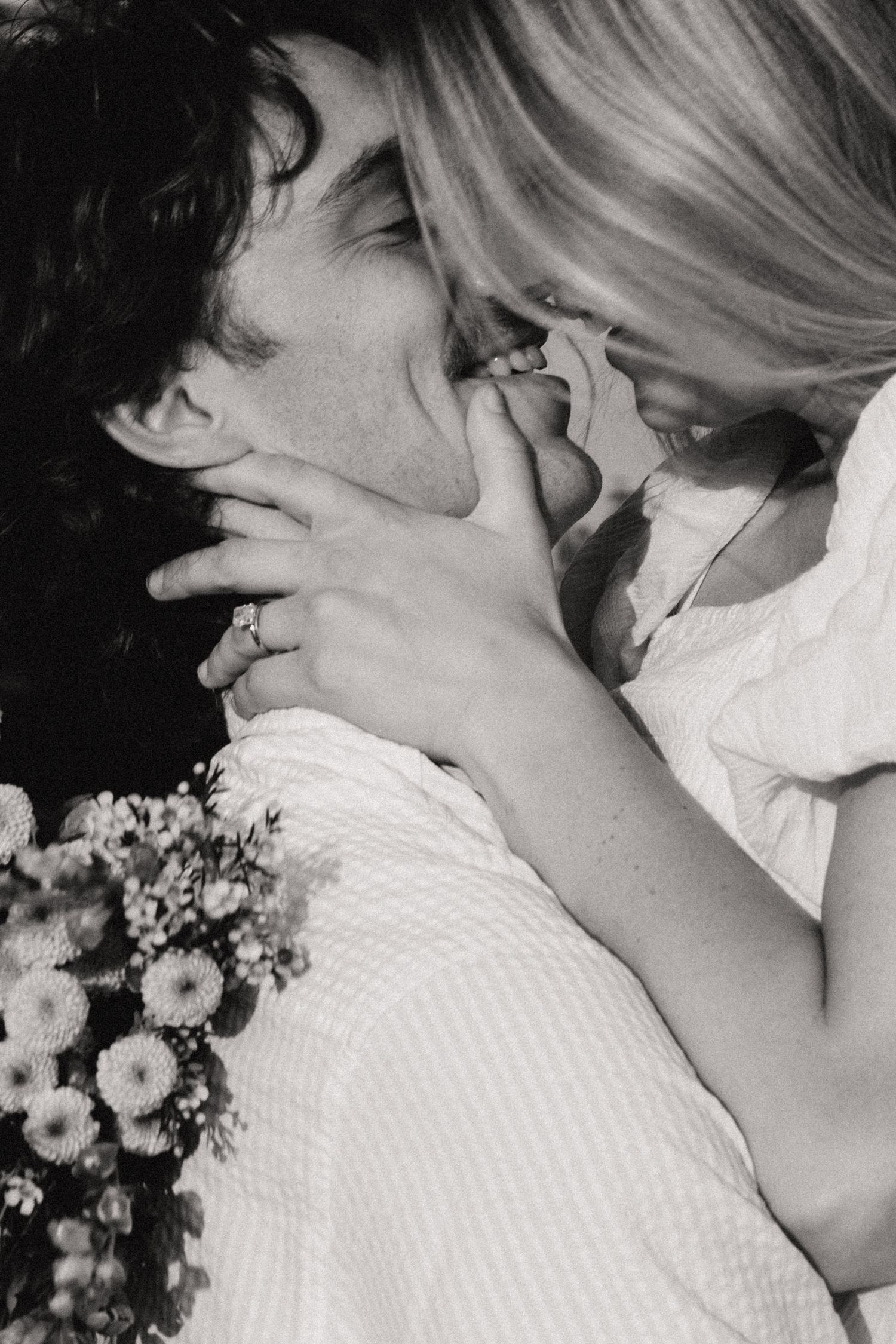 Black and white photo of a romantic couple kissing, woman holding bouquet of flowers, tender intimate moment.