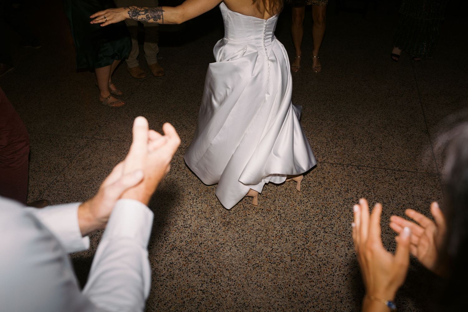 Bride in white wedding gown dancing at reception while guests clap in celebration.