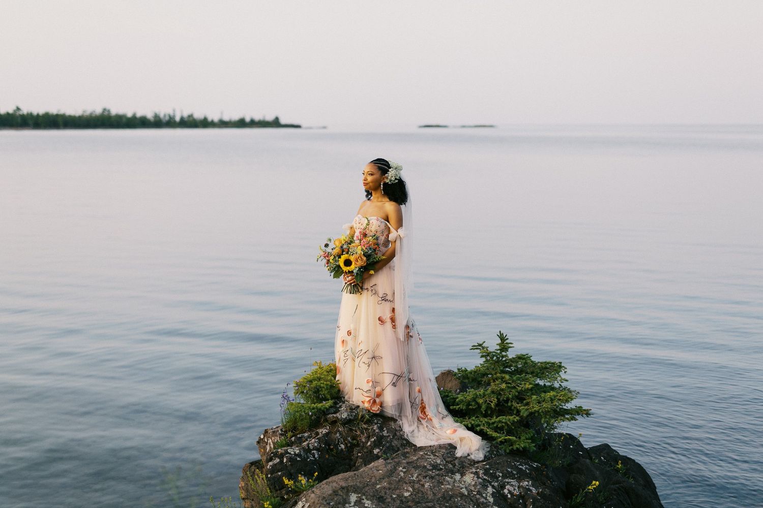 Bride in floral gown holds sunflower bouquet while standing on rocky shoreline at golden hour.