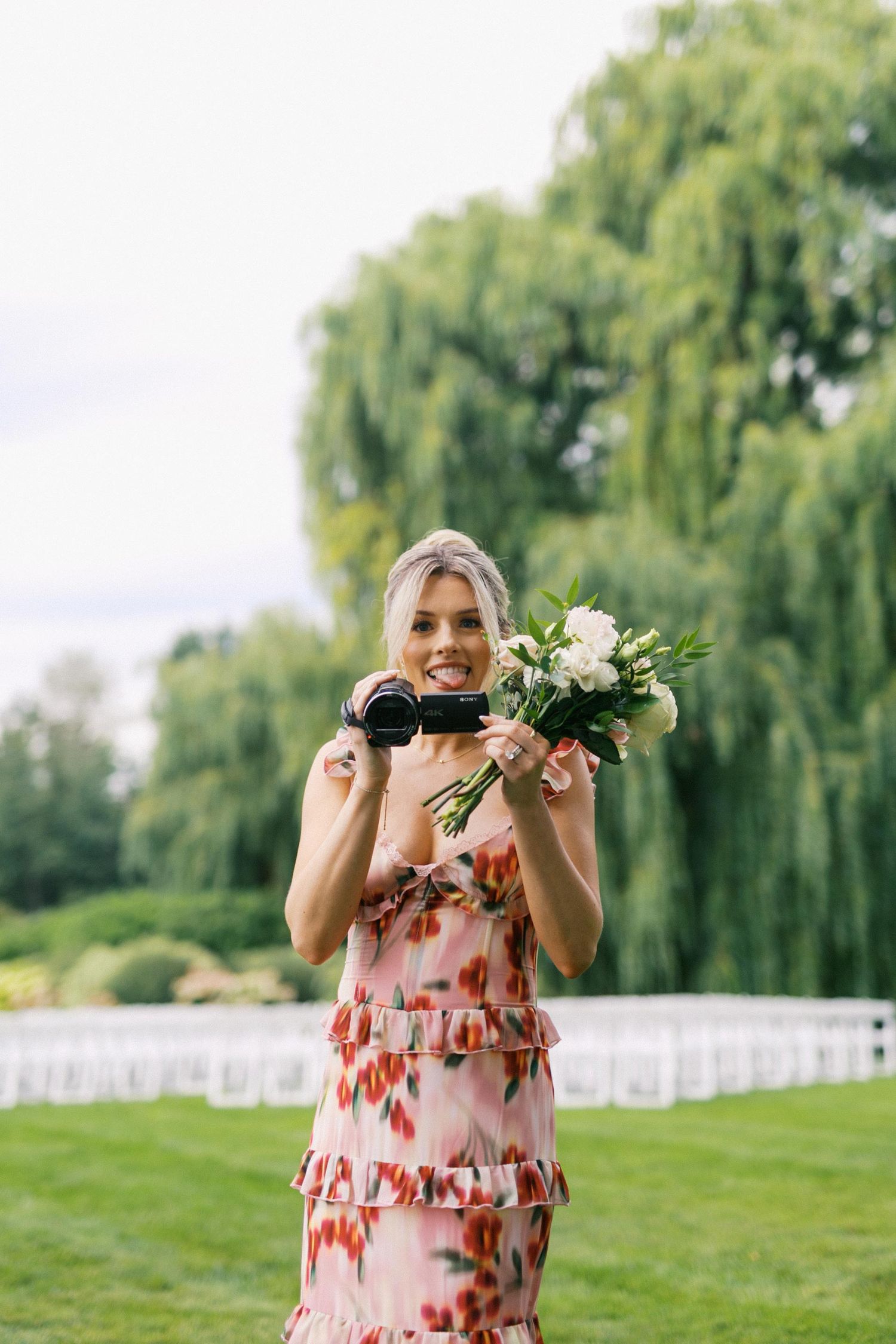 A bridesmaid in a pink floral dress holds up white flowers and video camera in front of their face in an outdoor garden setting.