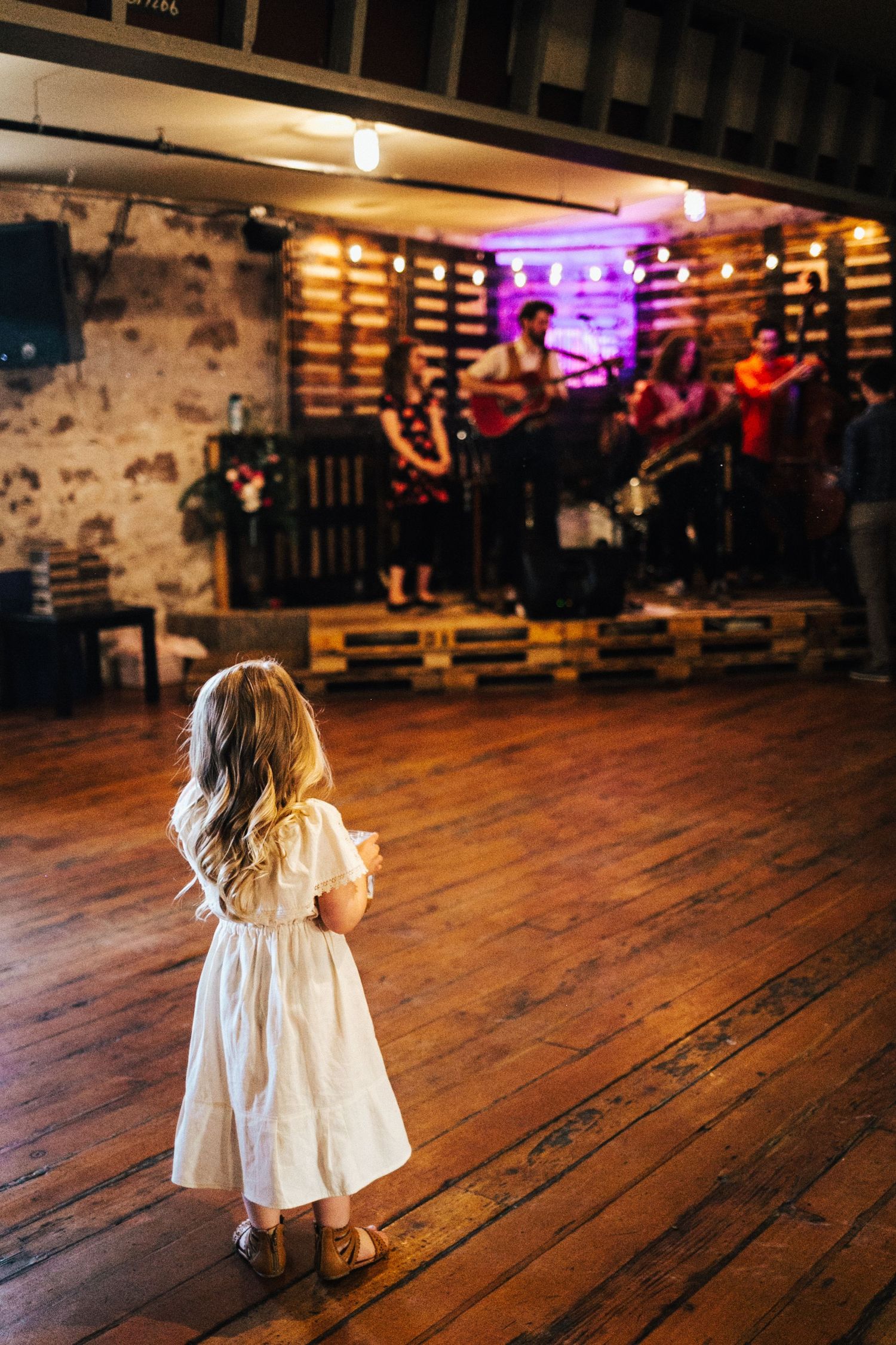 A small child in a white dress stands on a rustic wooden floor in a dimly lit venue with colorful lighting during wedding reception.