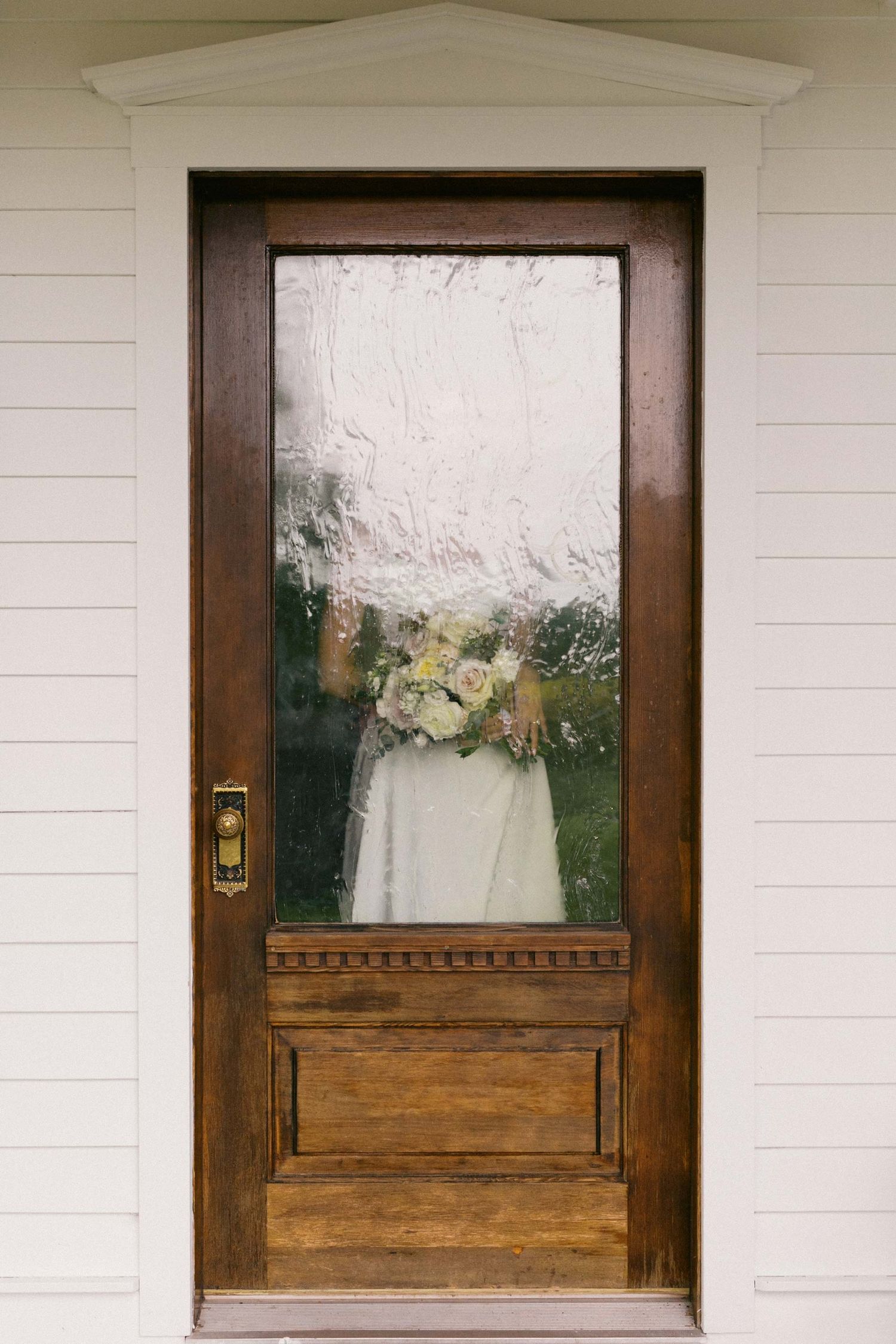A portrait of a bride and her bouquet through a wooden door with frosted glass panel.