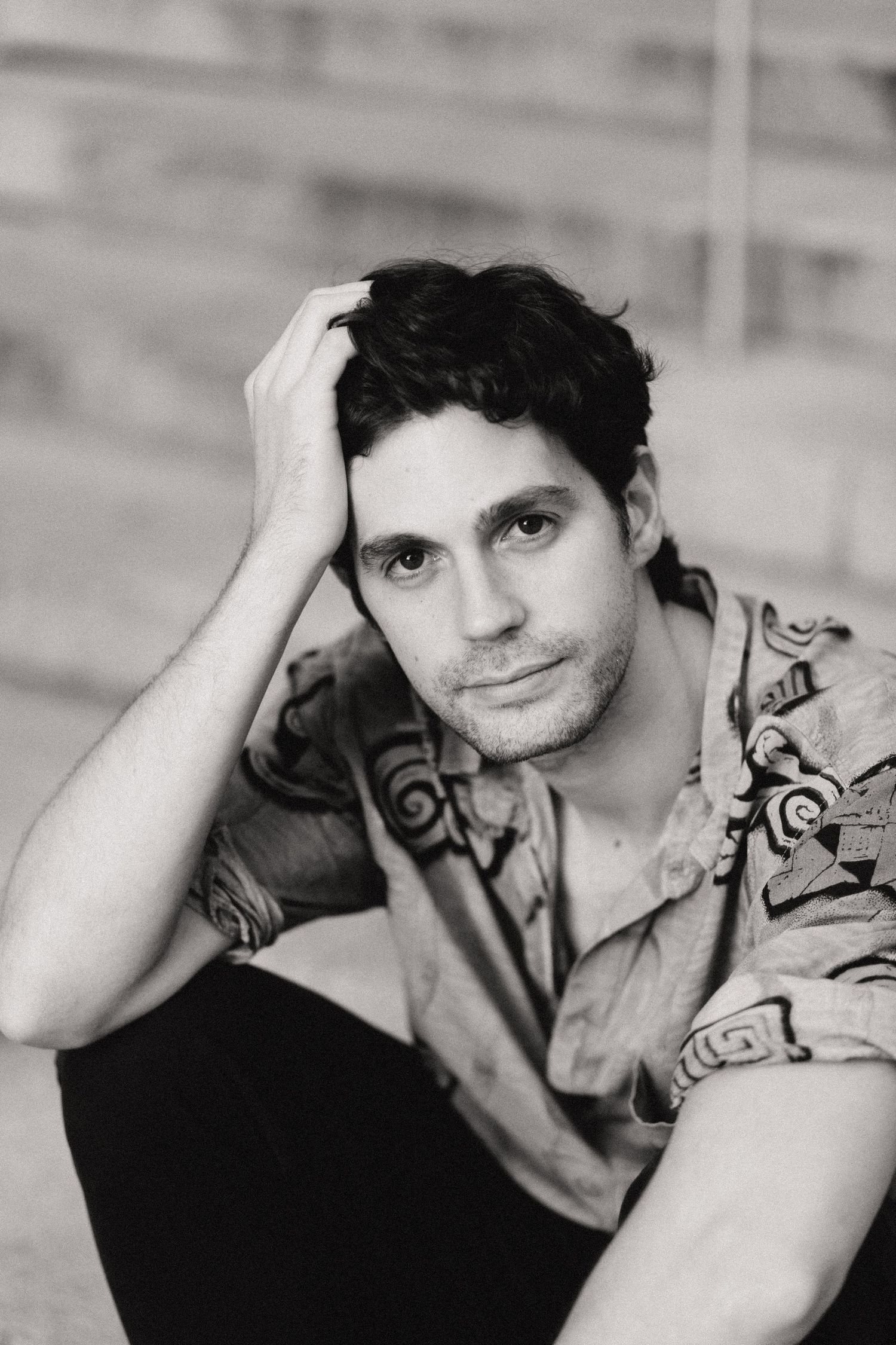 Black and white portrait of a handsome young man with curly hair sitting outdoors in a patterned shirt.