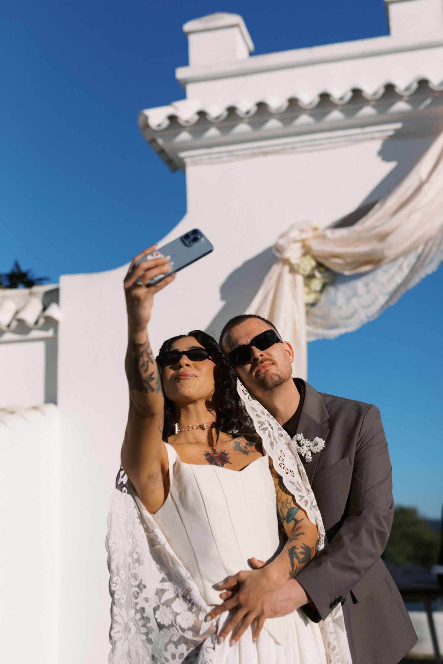 Tattooed bride and groom in sunglasses take a selfie outdoors at their wedding venue.