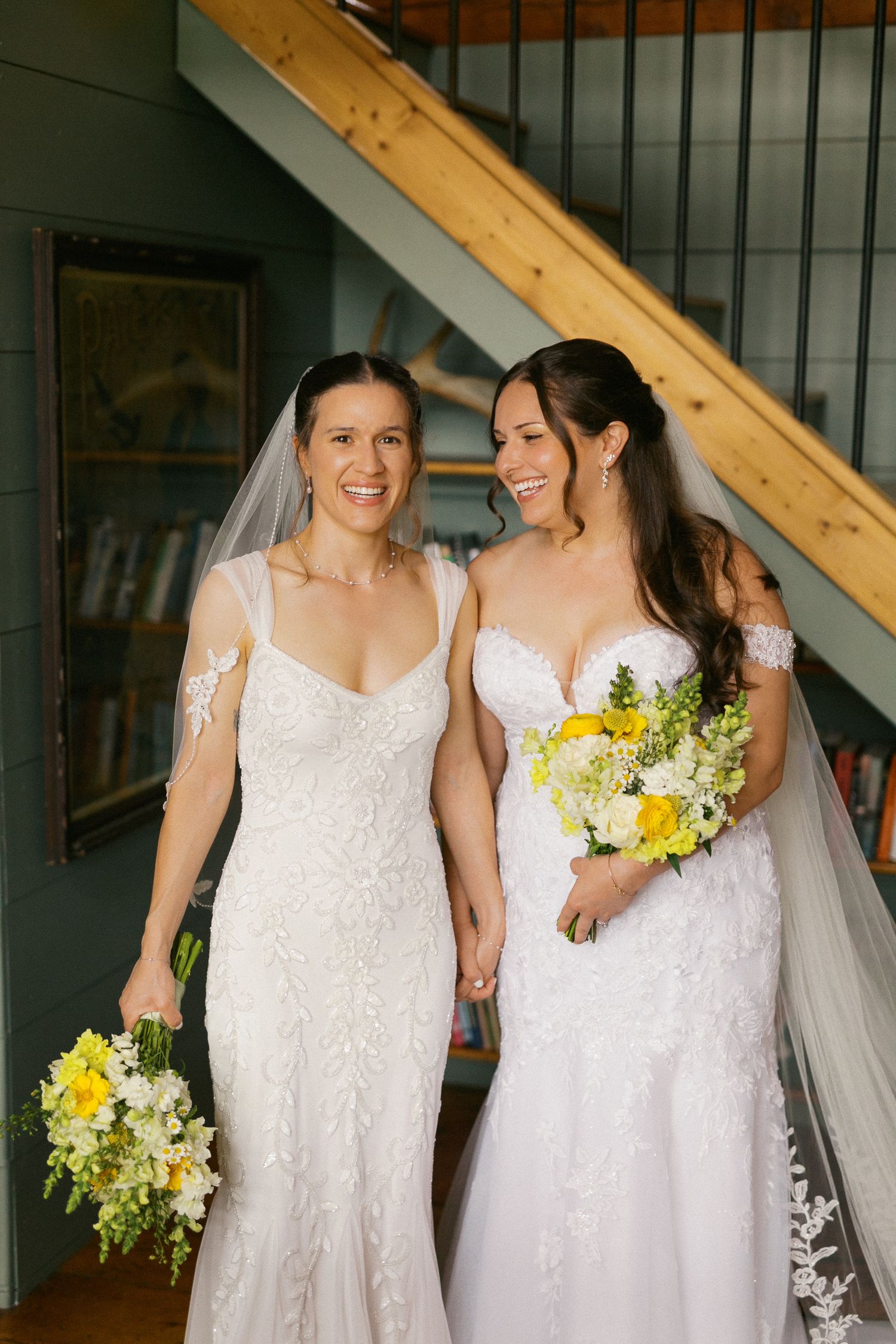 Two brides in white lace wedding gowns holding yellow bouquets and hands, smiling together near a staircase.