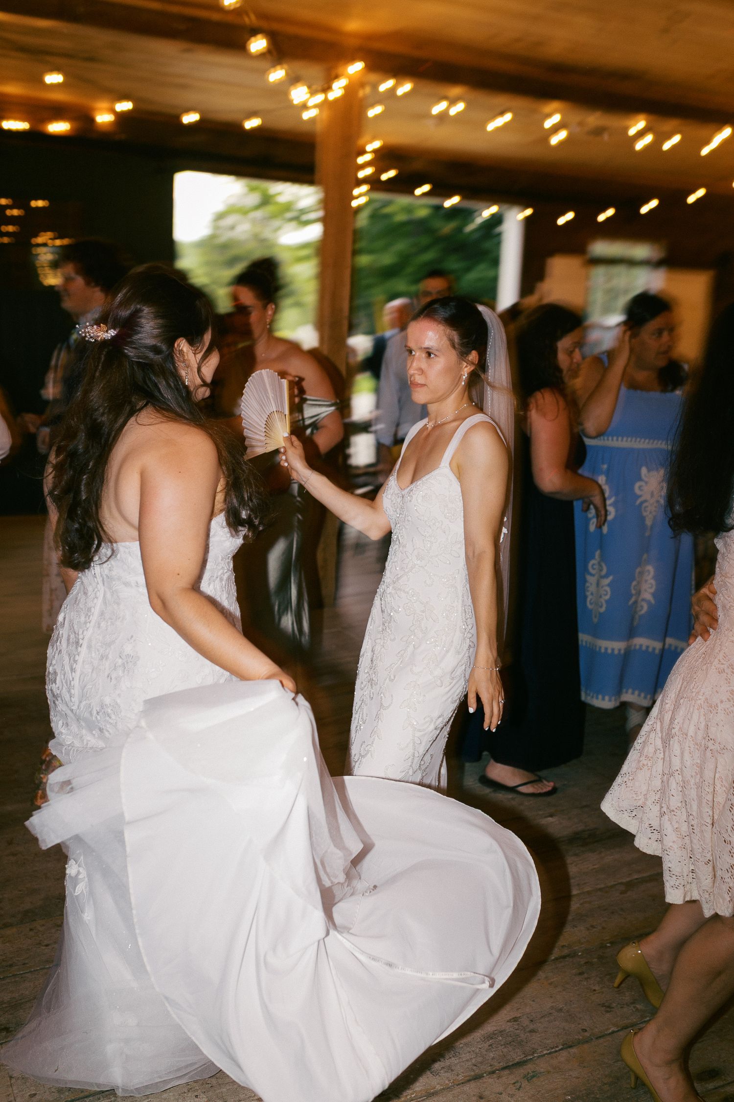 Two brides in white gowns dancing joyfully at their wedding reception in a rustic venue with string lights.