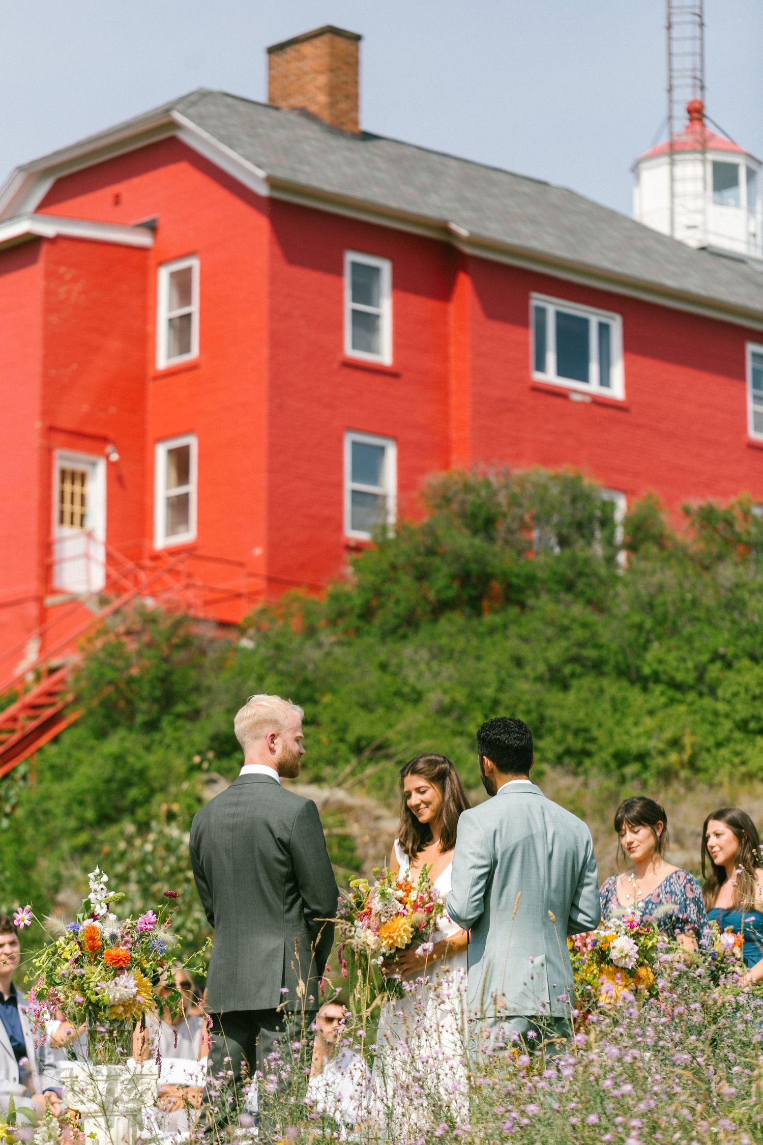 Wedding guests gather in front of a bright red lighthouse building surrounded by greenery.