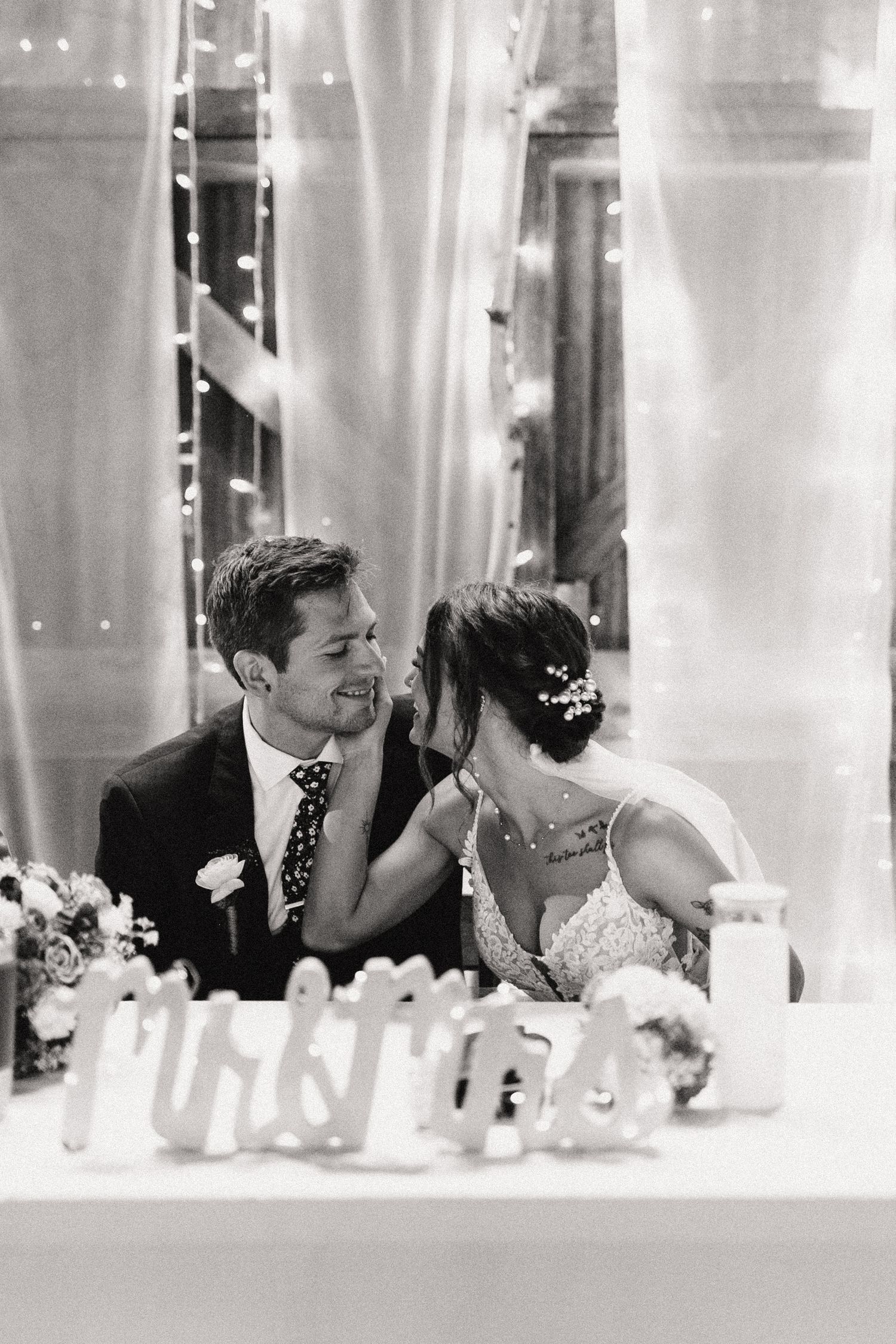 A tender black and white candid moment between newlyweds at their reception table with soft lighting and white decor.