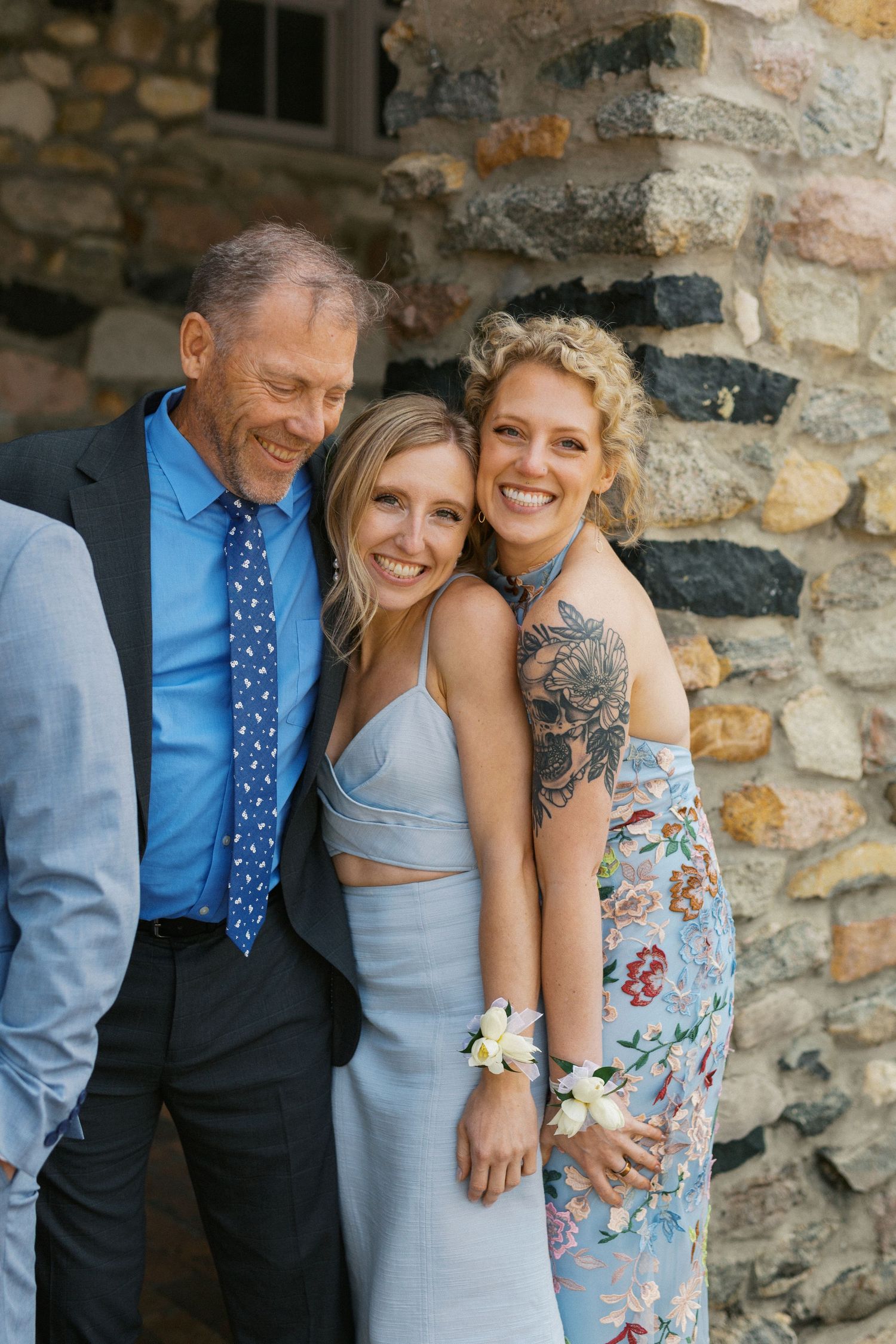Three people share a warm embrace against a rustic stone wall background during a joyful outdoor celebration.