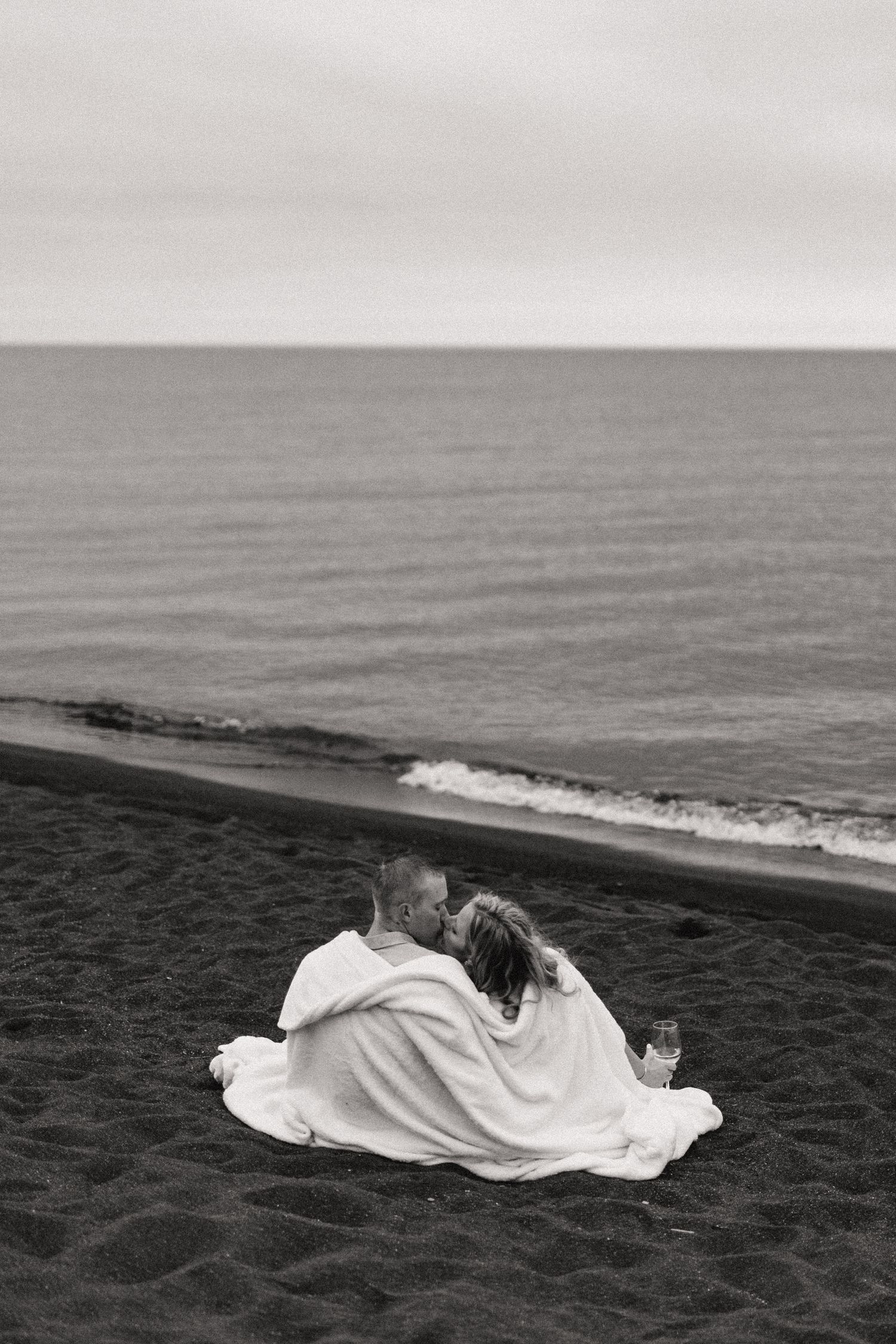 A romantic couple wrapped in a blanket sits together overlooking the ocean on a cloudy day.