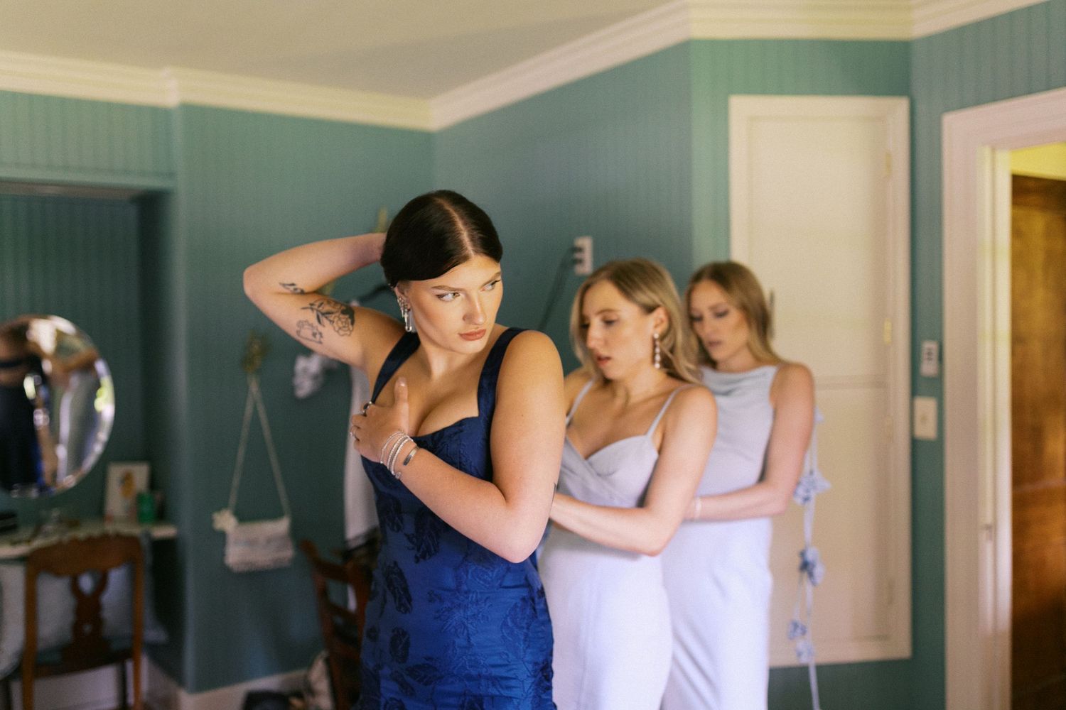 Bridesmaids in blue dresses help with wedding preparations in teal-walled room.
