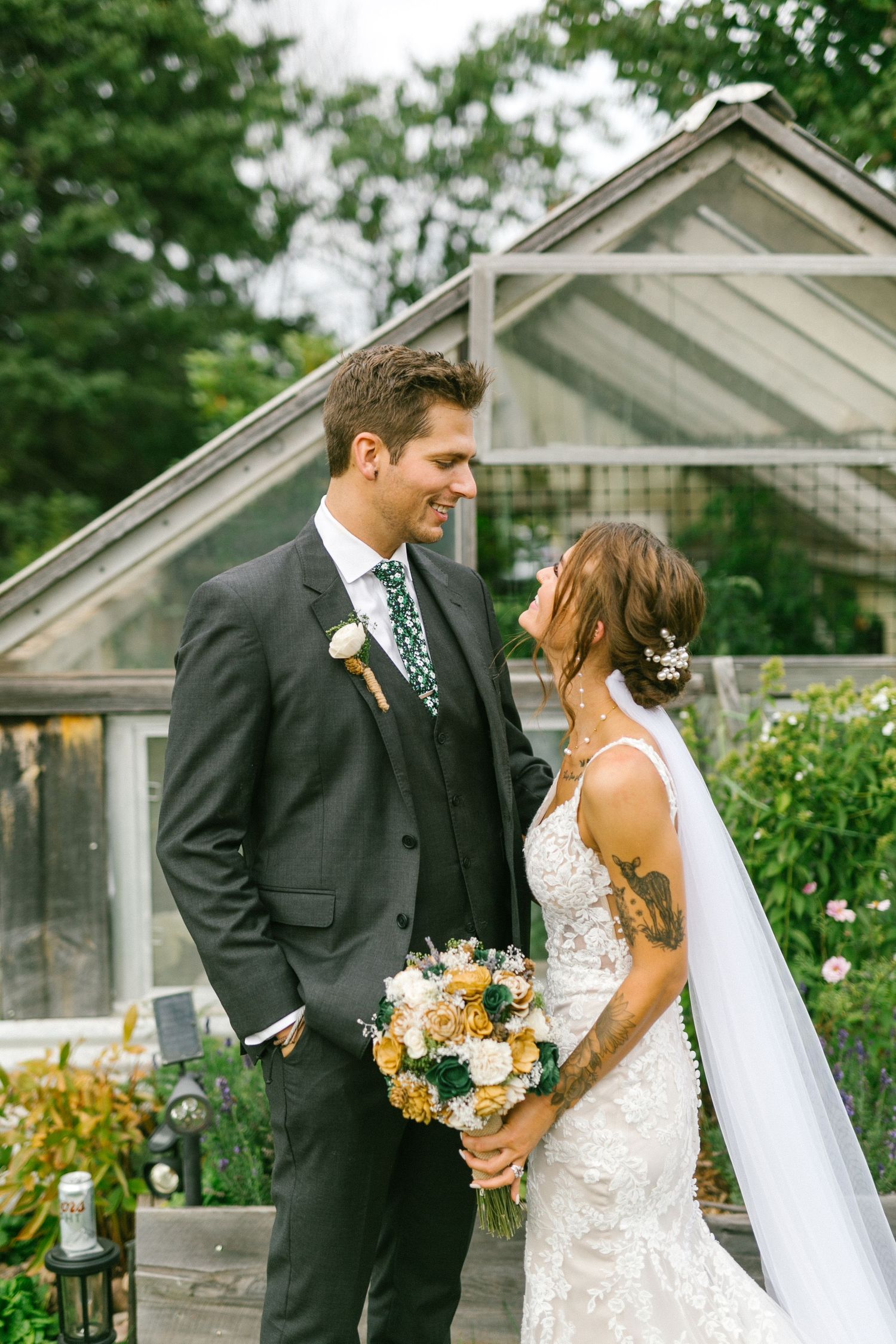 A bride and groom share an intimate moment together in front of a greenhouse surrounded by lush gardens.