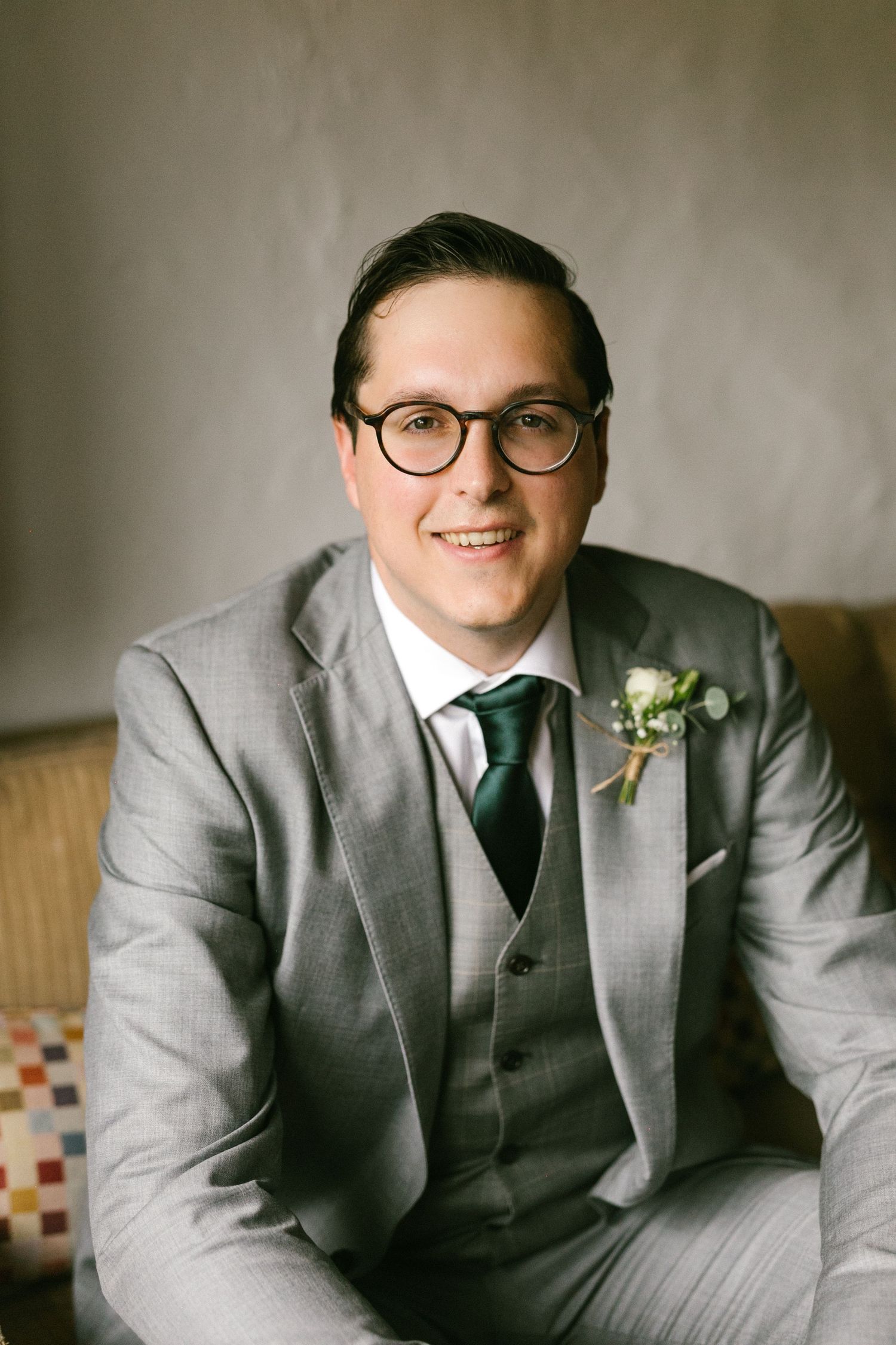 El novio lleva un traje clásico gris de tres piezas con corbata verde y boutonniere sentado en un mueble tapizado.