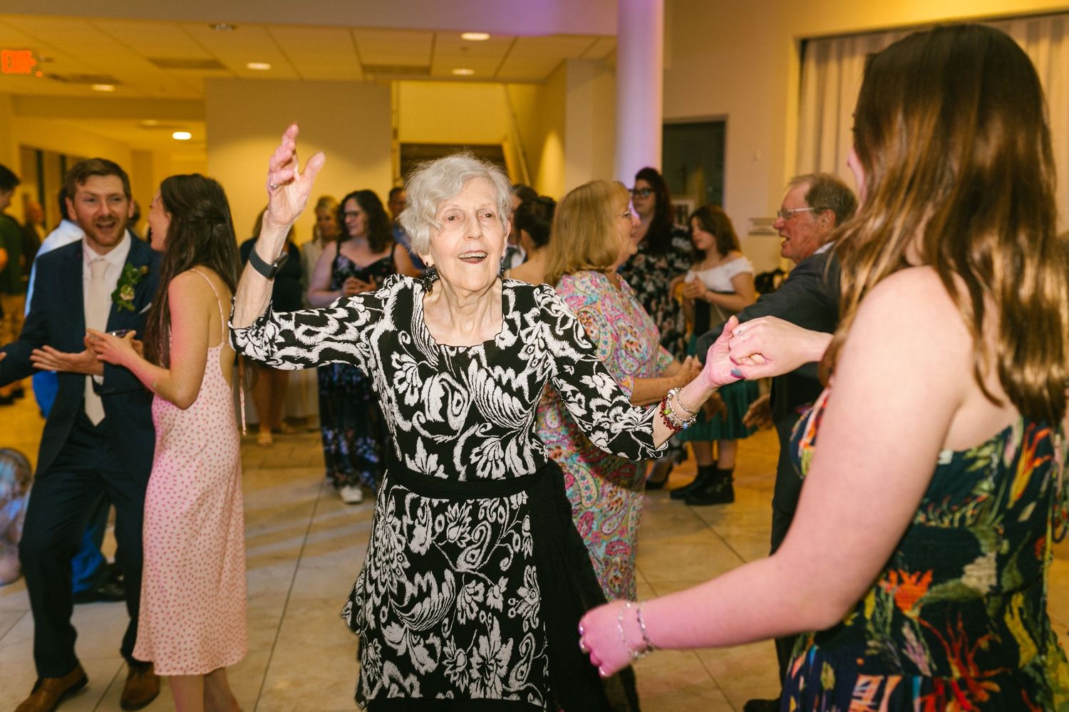 An elderly woman dances joyfully in a black and white patterned dress at a lively indoor celebration.