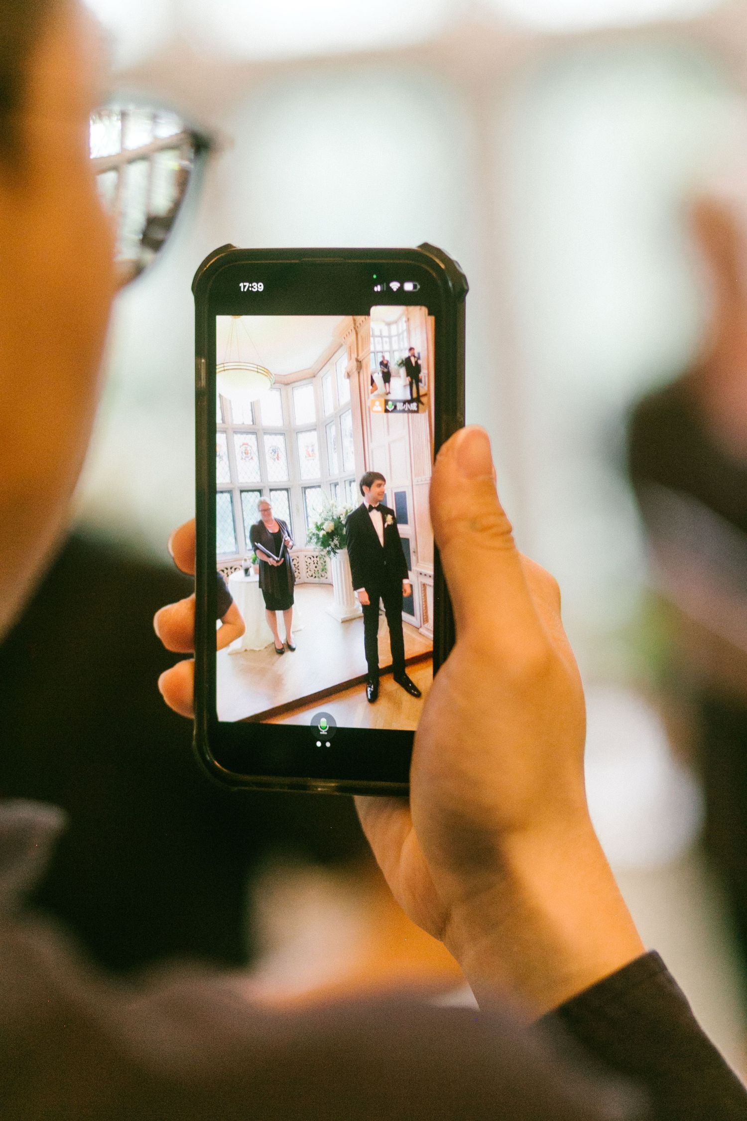 Wedding guest photographs the groom and a woman in a bright sunlit room on a smartphone.