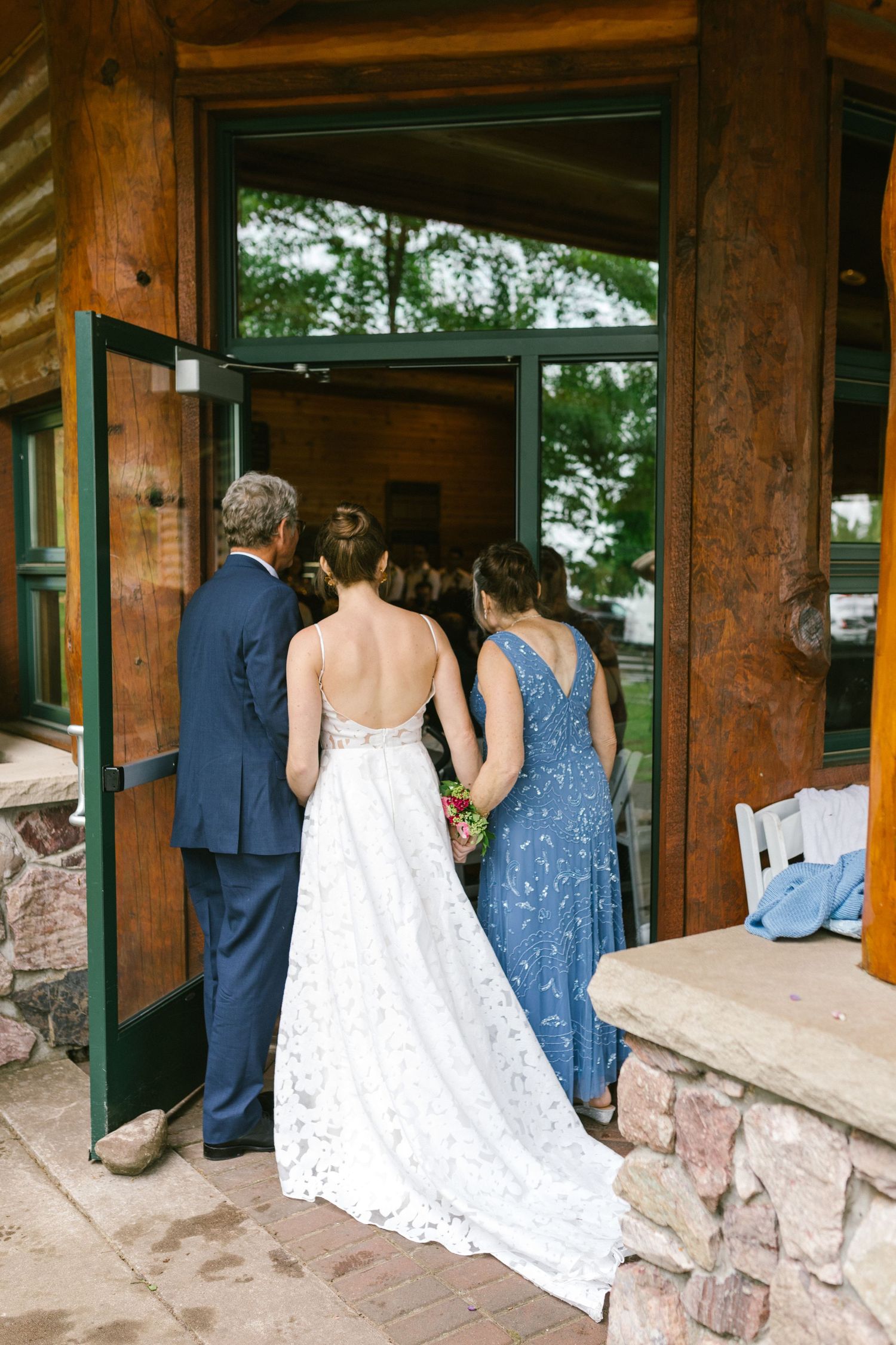 Wedding party enters rustic lodge building with stone foundation and wooden beams, bride wearing flowing lace gown.